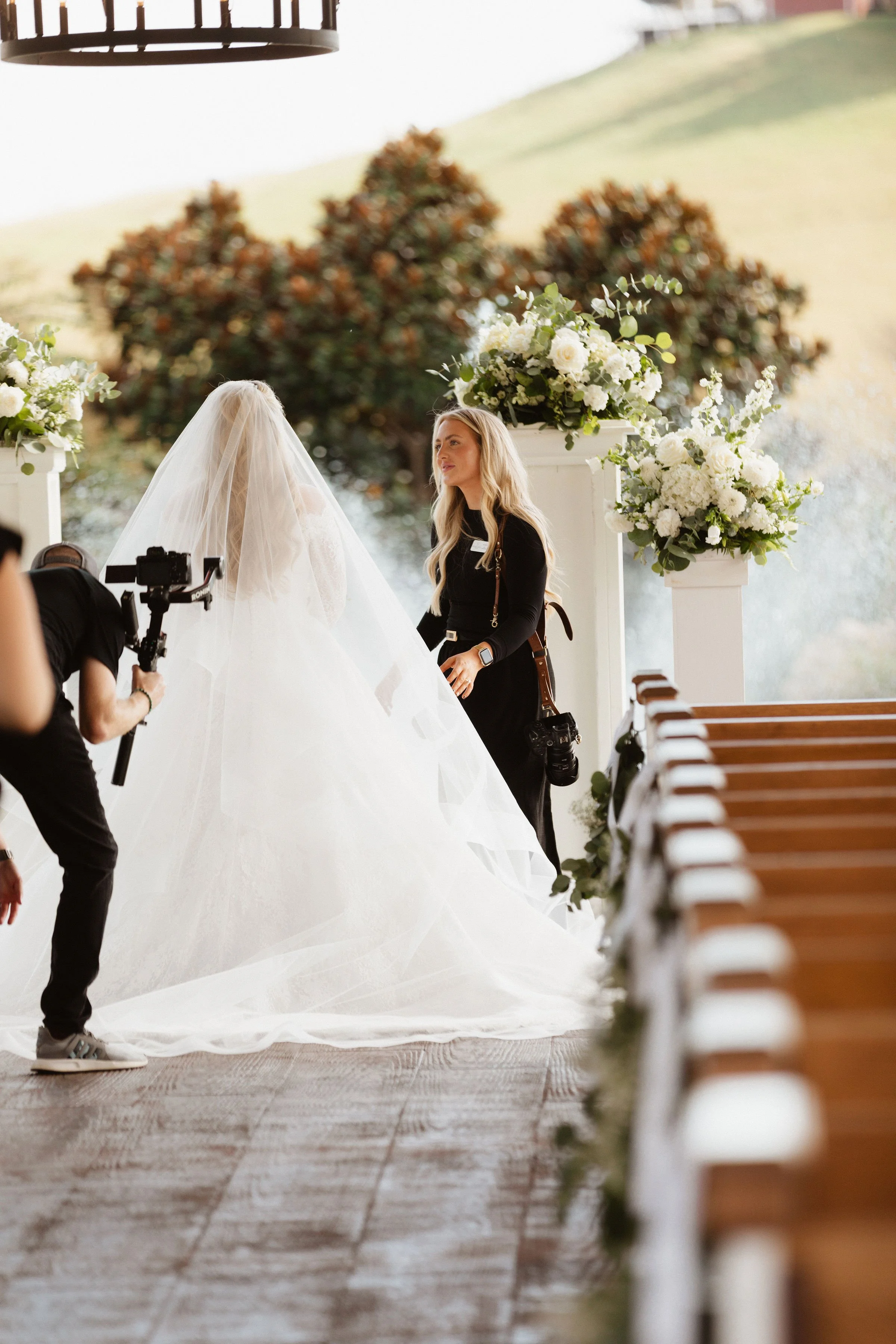 A bride in a white wedding gown and veil stands in front of a woman during a wedding ceremony outdoors, under a canopy of trees, with floral arrangements and empty pews along the aisle.
