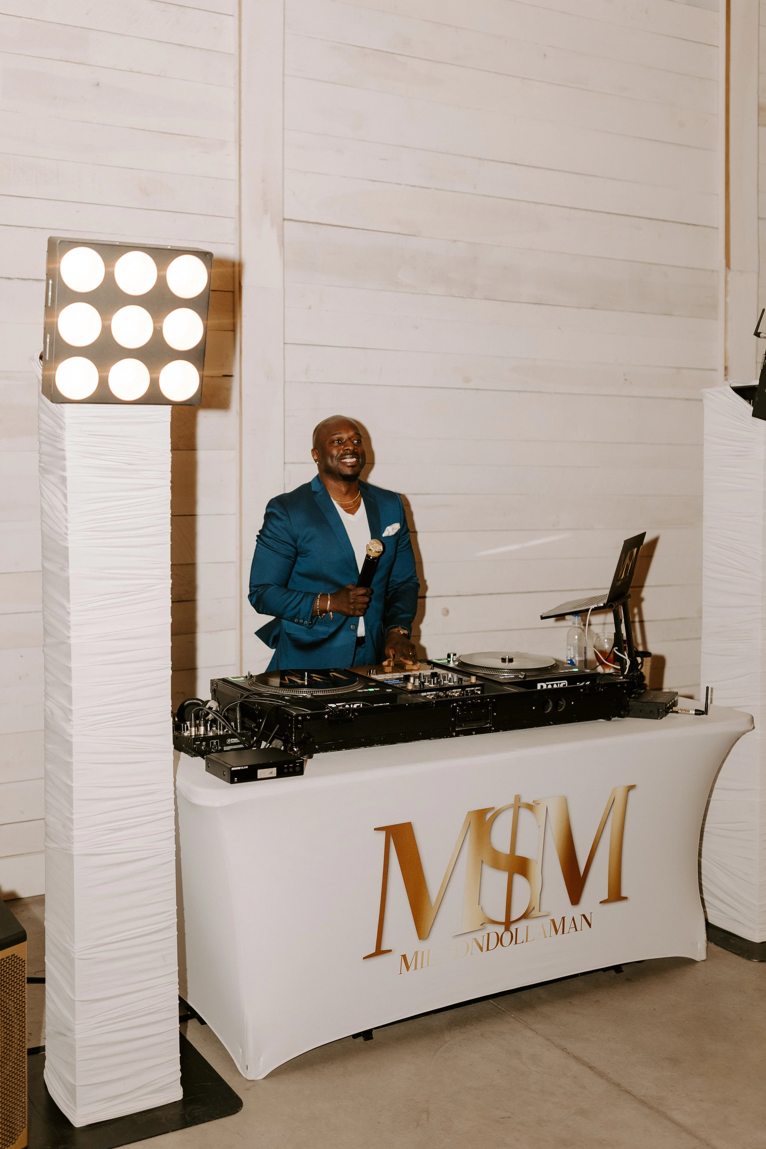 A man in a blue suit standing behind a DJ table with turntables and a laptop, holding a microphone, at an event with white decor.