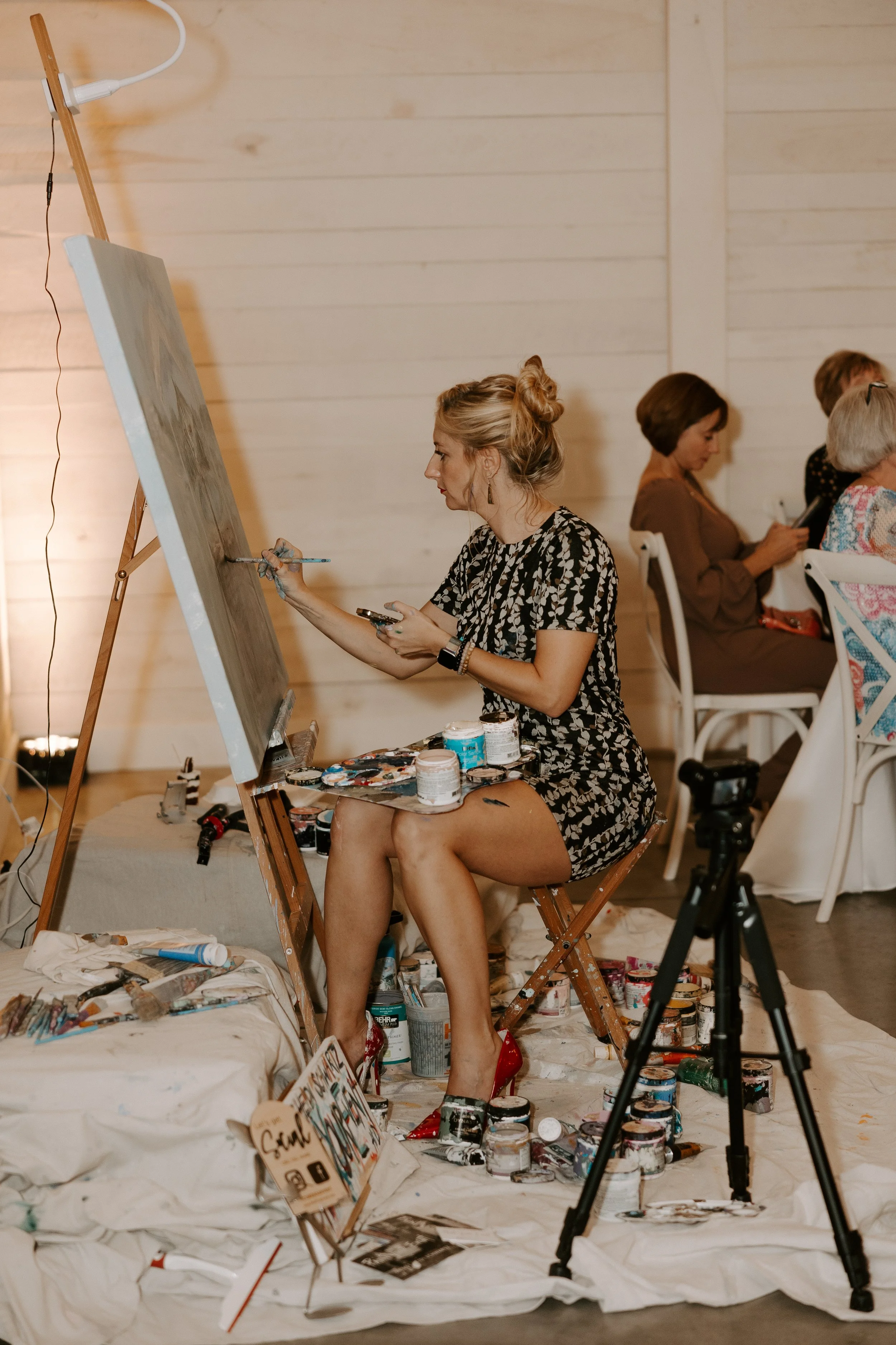 A woman in a black and white dress painting on a large canvas at an art event. She is sitting on a wooden chair, surrounded by paint cans and brushes, with her feet in red high heels. Other seated women are in the background.