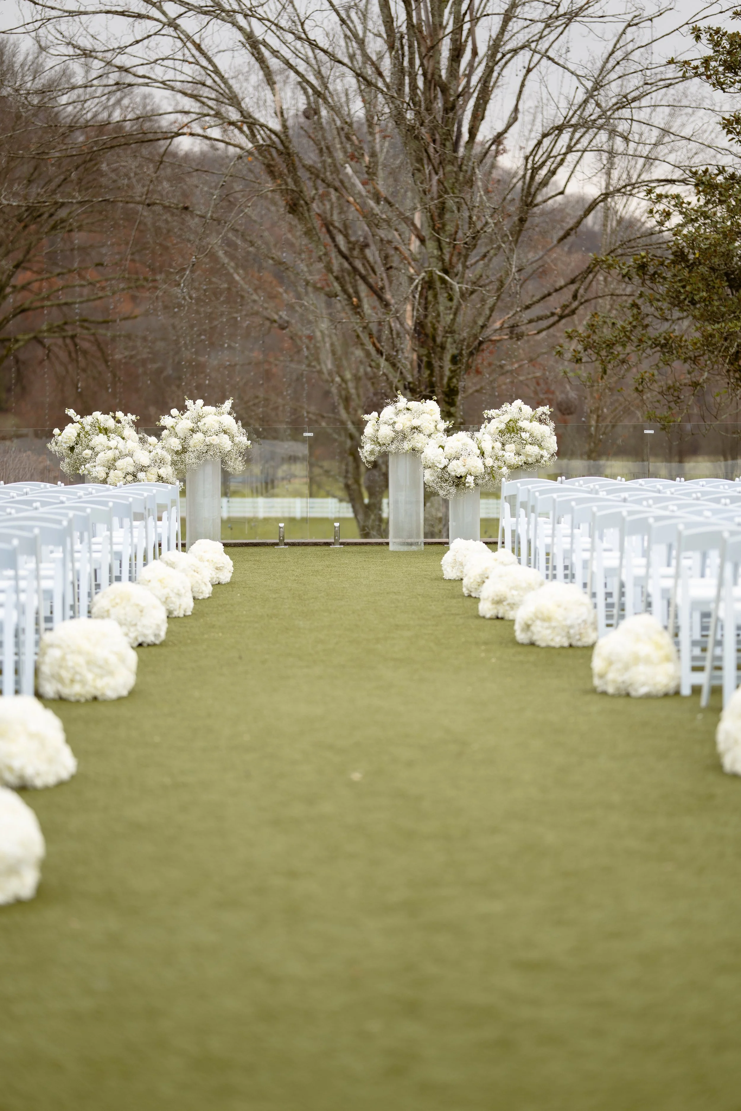Empty outdoor wedding aisle decorated with white flowers leading to a floral arch in front of a large bare tree, set against a cloudy sky.