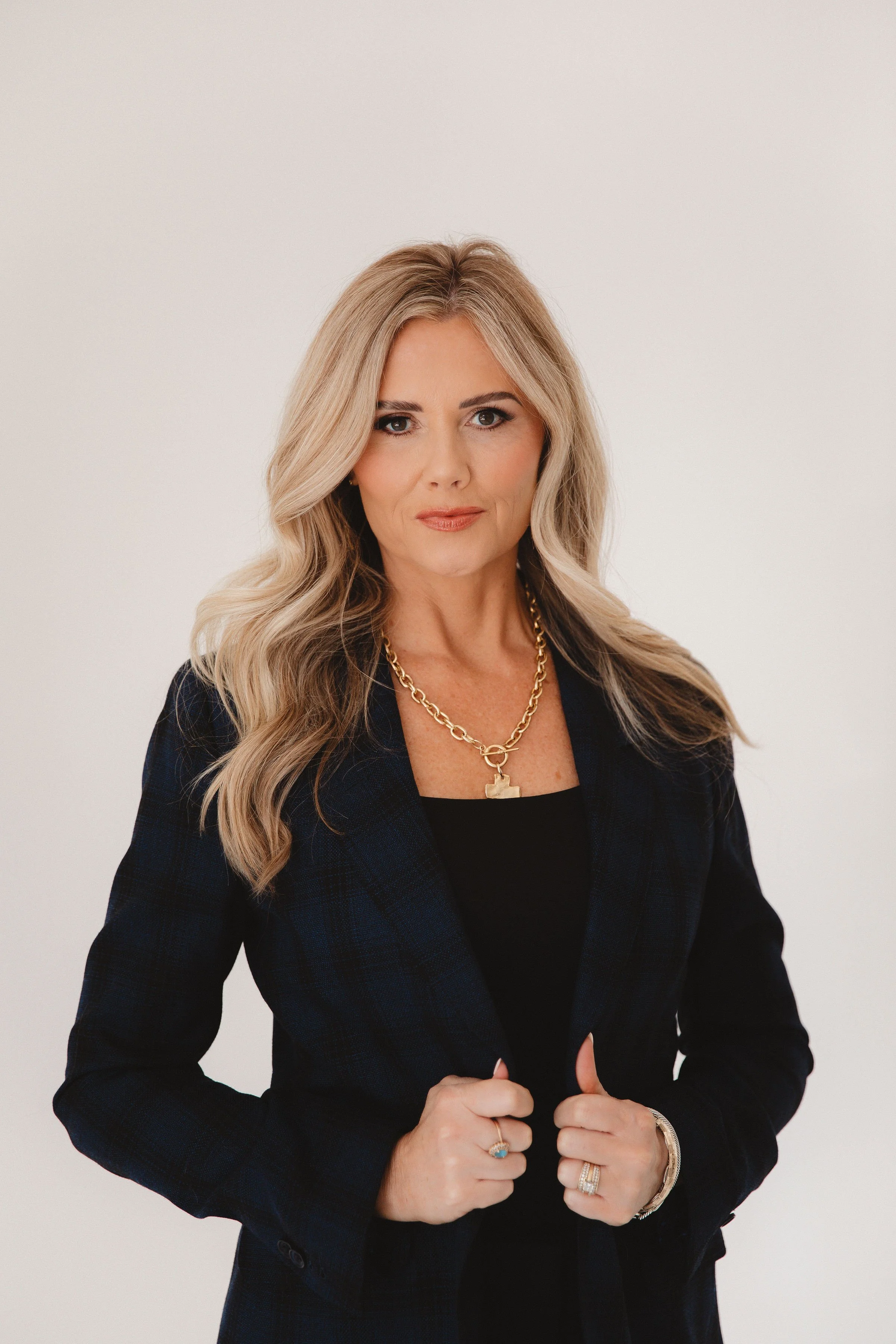 A woman with long blonde hair, smiling, wearing a sleeveless dark denim vest with gold buttons and a gold chain necklace, against a plain light background.
