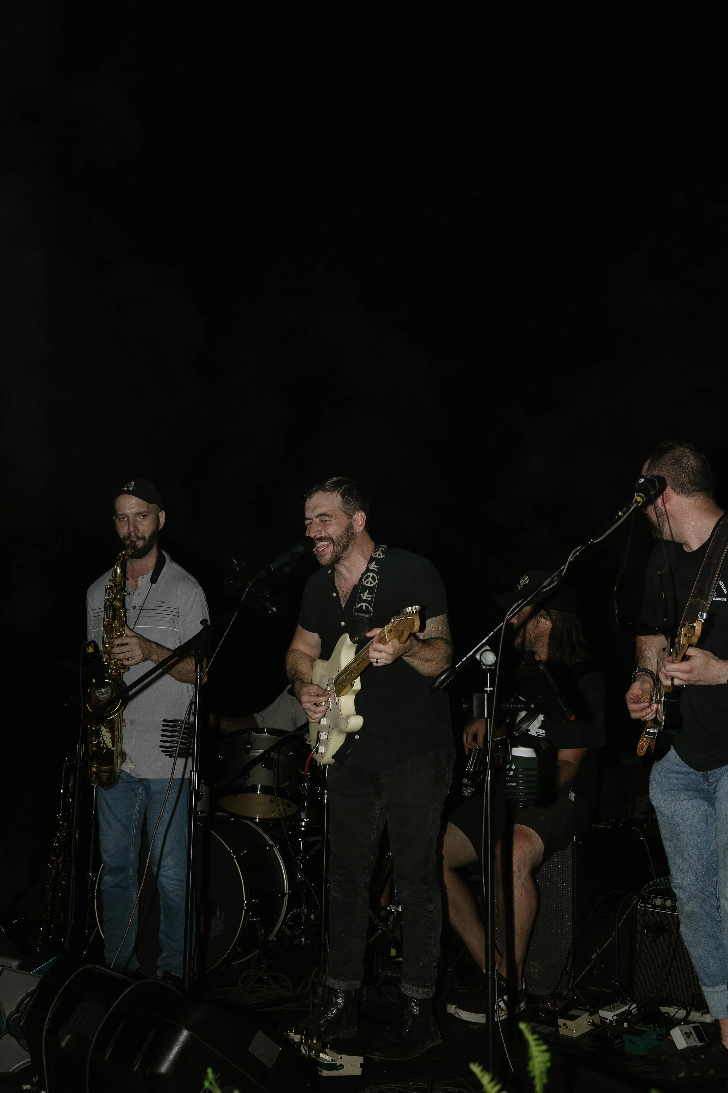 A band performing on stage, with a guitarist singing into a microphone, a saxophonist, and a bassist, all in a dark setting.