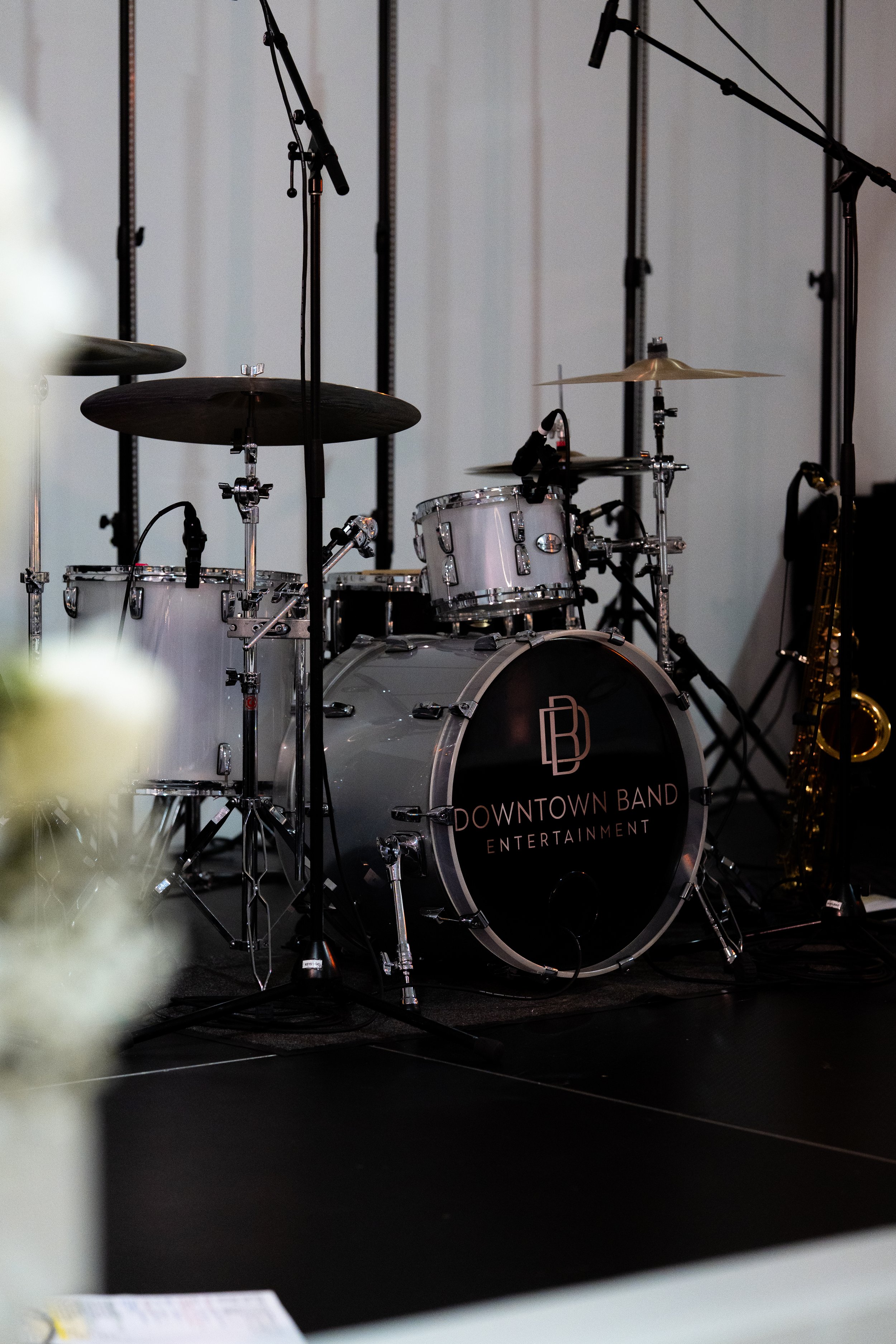 A set of white drum kit with a bass drum labeled 'DOWNTOWN BAND ENTERTAINMENT', surrounded by microphone stands, in a performance stage setting with saxophone visible to the right.