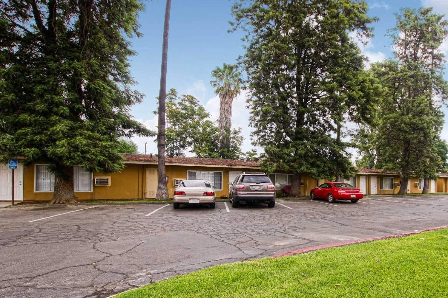 This image features a single-story apartment complex with mustard-yellow walls and front-row parking. The landscape includes lush trees. 