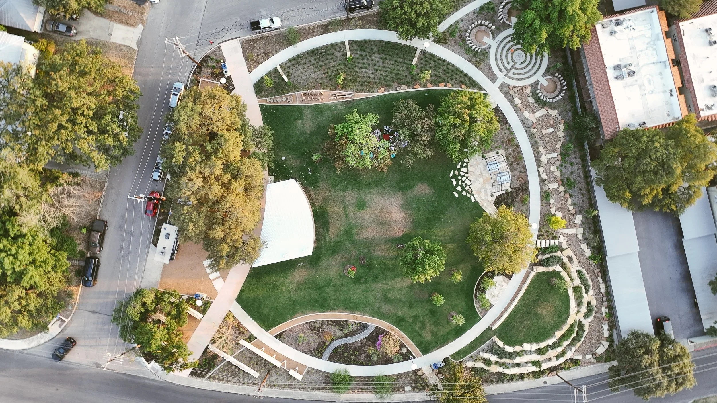 An aerial view of a park with green grass, trees, and paths forming a spiral and circular patterns. Surrounding the park are streets with parked cars and nearby buildings.
