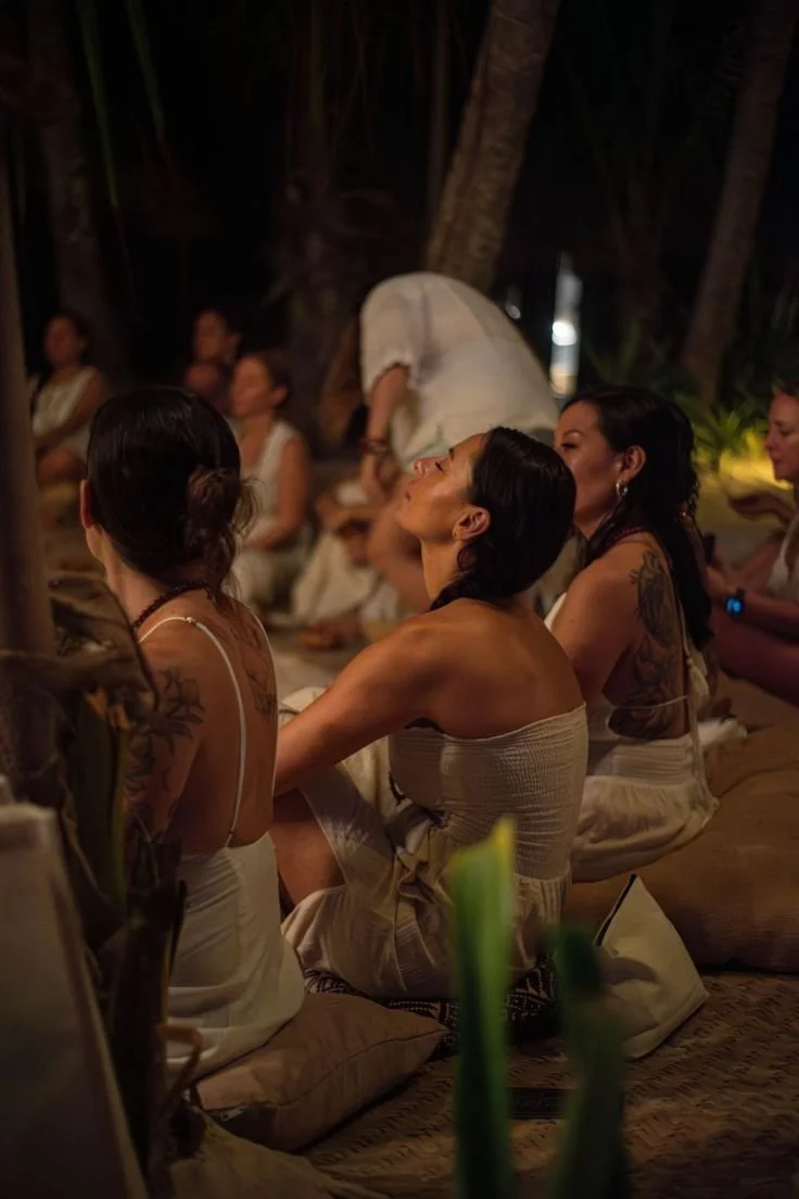 Group of women sitting on cushions outdoors at night, some with visible tattoos, in a tropical setting with trees.