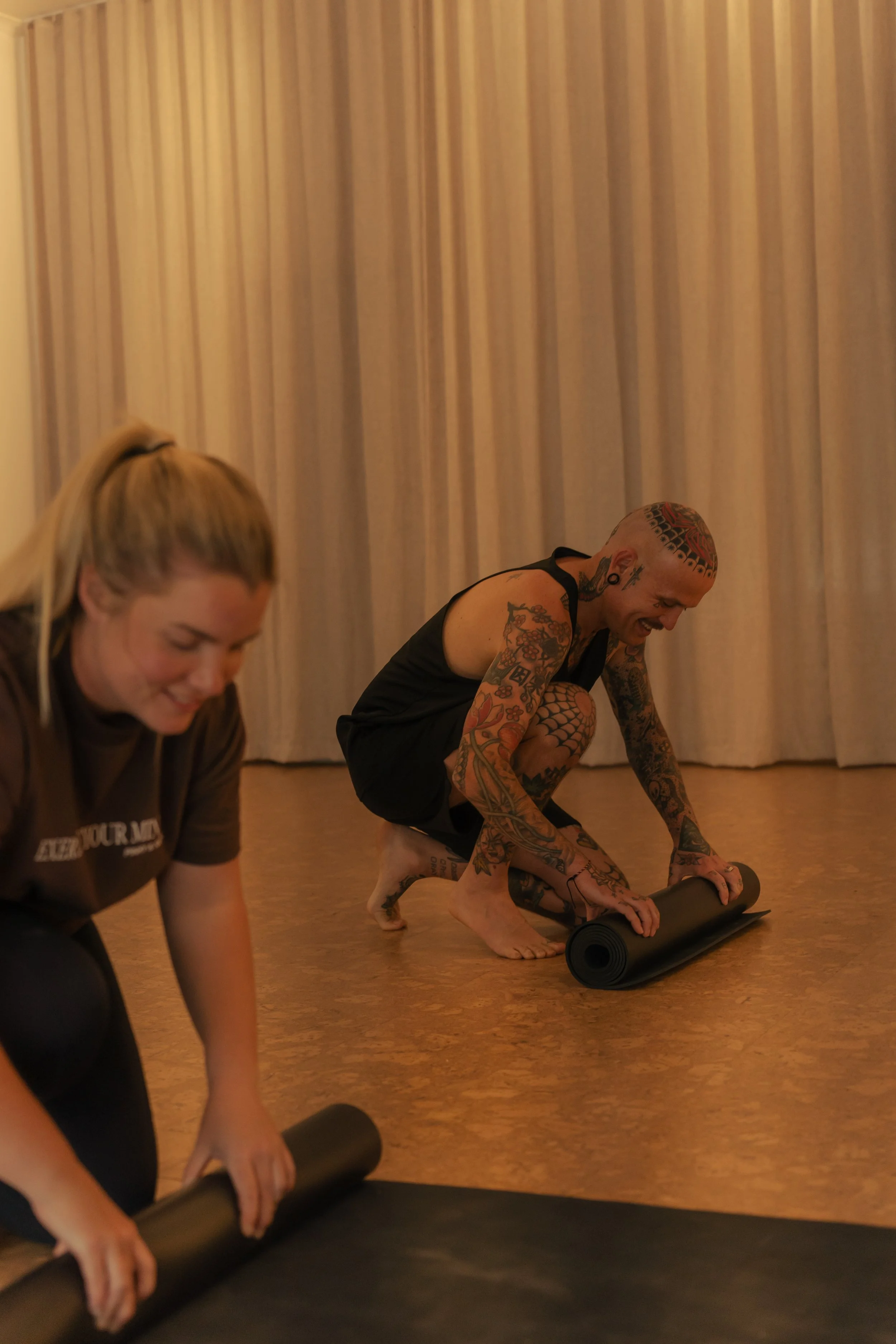 Two people with tattoos, one female and one male, engaging in a yoga or stretching exercise on a wooden floor with beige curtains in the background. They are lifting foam rollers, possibly in a fitness or yoga class.