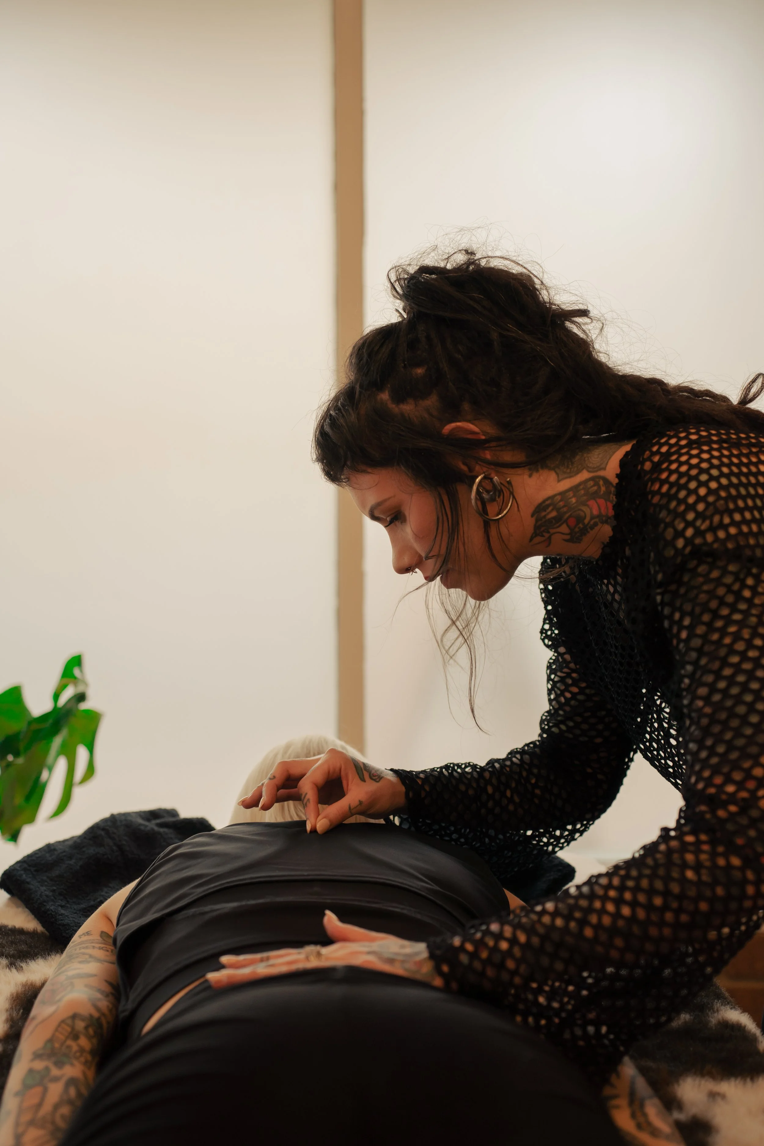 A person with dark, curly hair and tattoos on their neck and hand is receiving a massage or similar treatment from another person with tattoos on their arm, in a room with beige walls and a small green plant.