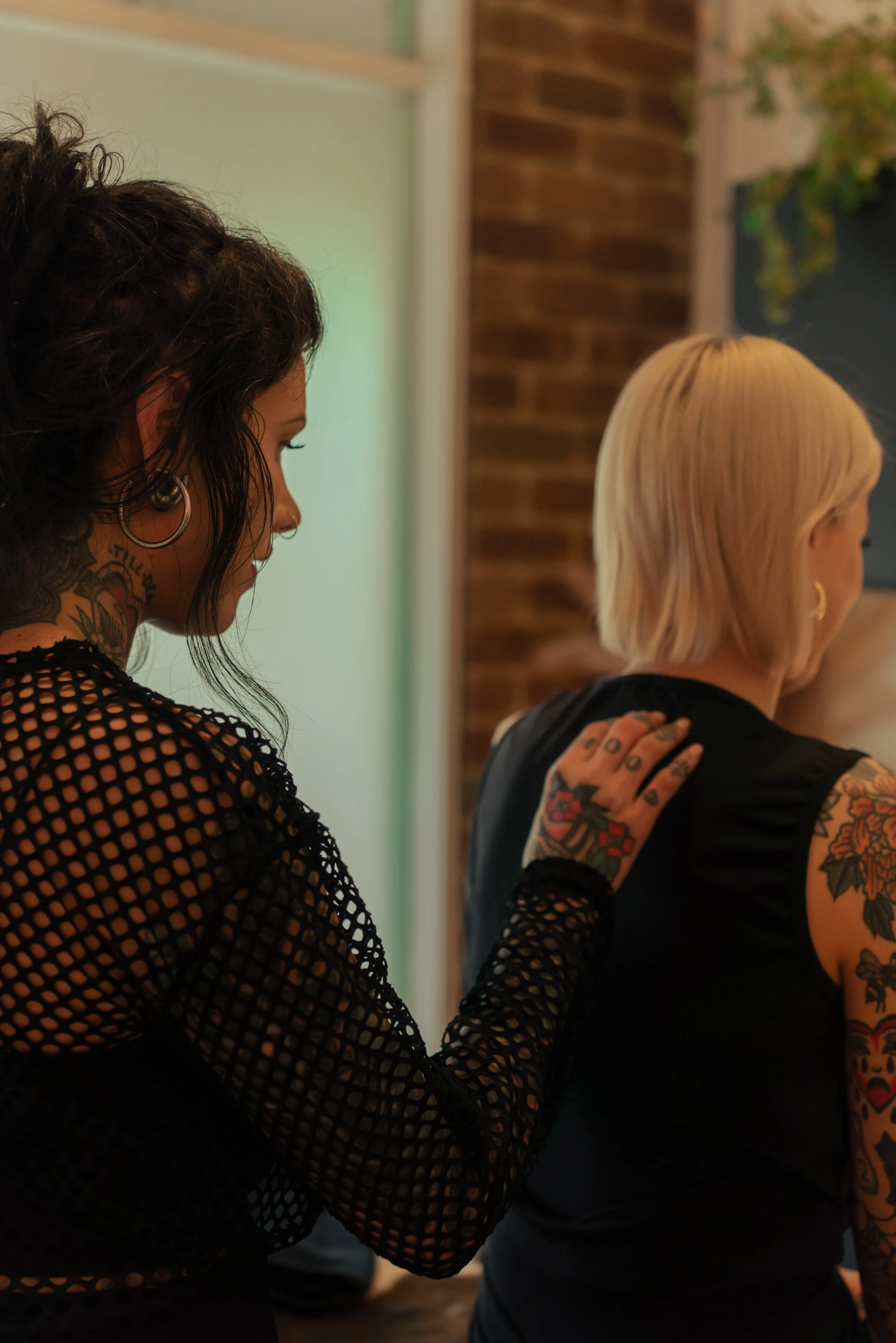 Two women with tattoos standing close together in an indoor space, one with dark hair and the other with blonde hair, both wearing black attire, against a background with a brick wall and a plant.