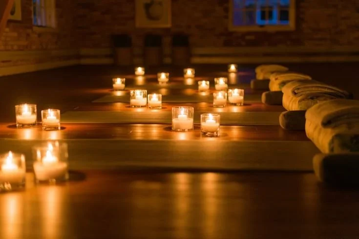 A dimly lit room with multiple lit candles on a long wooden table, surrounded by cushioned seating.