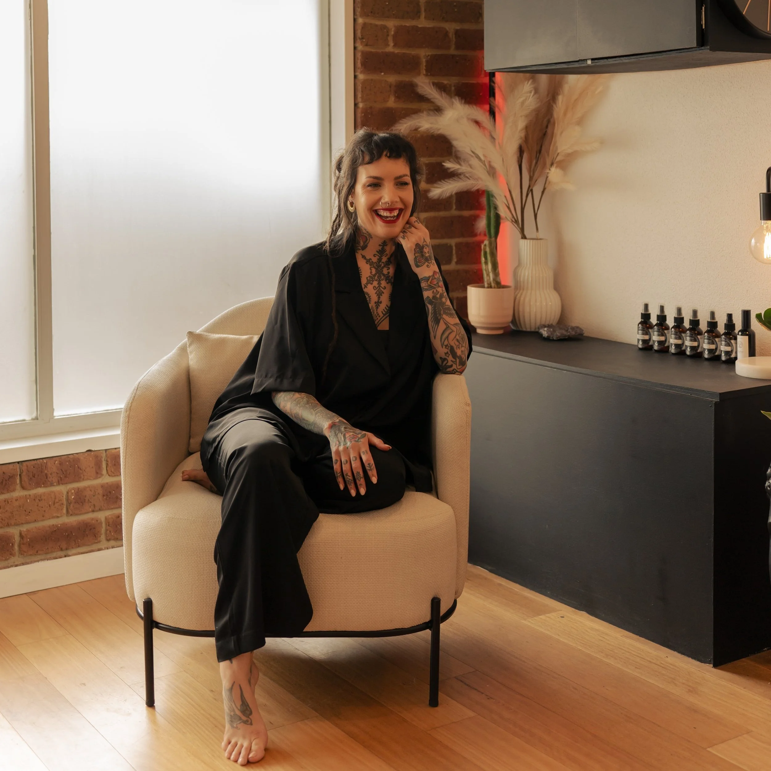 A woman with tattoos and dark hair, wearing black clothing, smiling while sitting on a beige armchair in a room with brick and white walls, next to a black cabinet with decorative items.