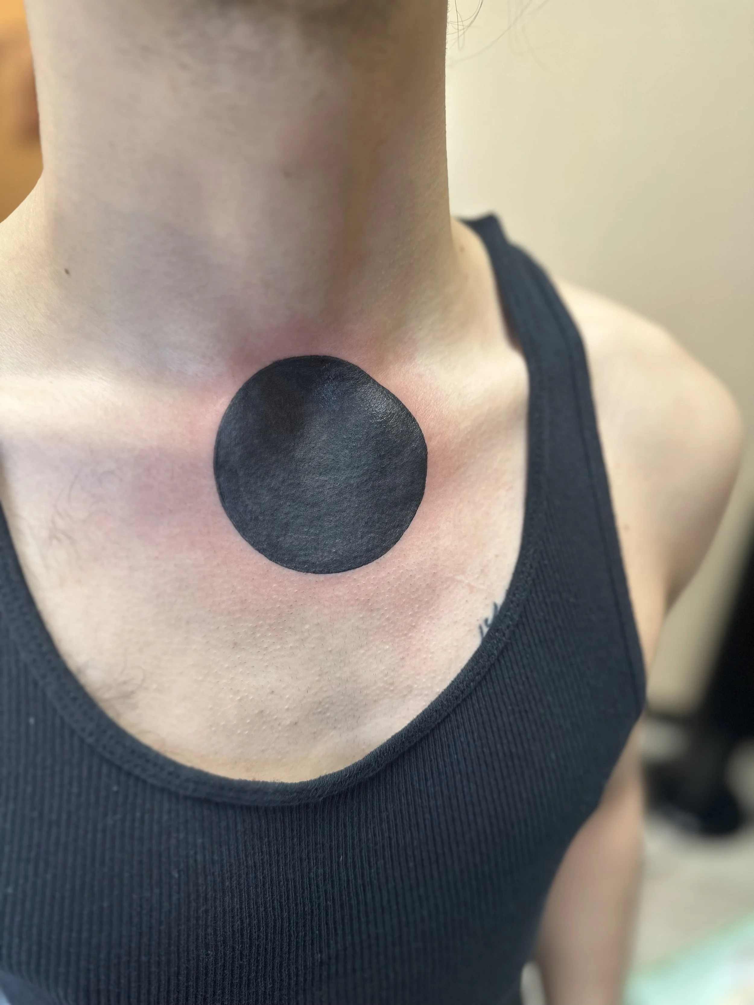 A person with a black circle tattooed on their lower neck, above the collarbone, wearing a black sleeveless top.