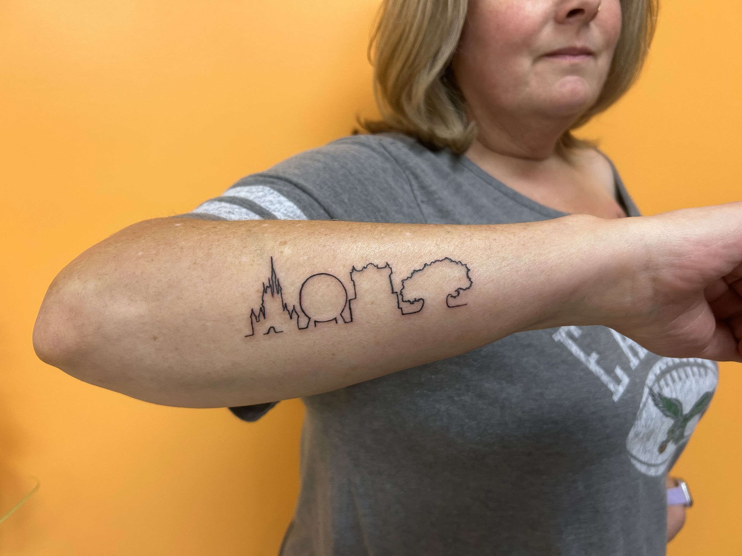 A woman with blonde hair shows a tattoo on her arm of the word 'LOVE' with skyline and cloud illustrations. its the Disney castles!