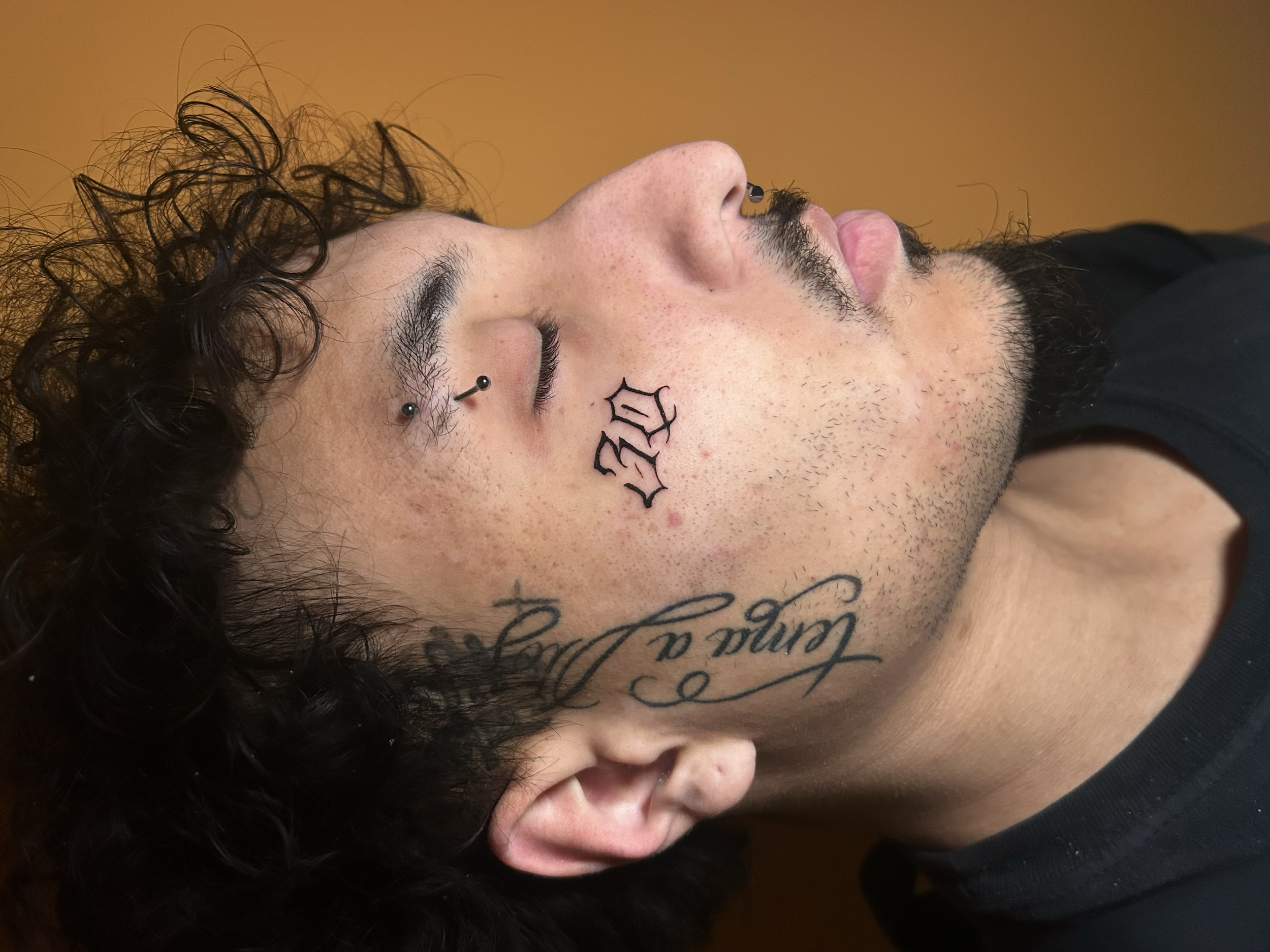 Close-up side profile of a person's face with dark curly hair, skin freckles, tattoos on the cheek and neck, a nose piercing, and a mustache and beard.