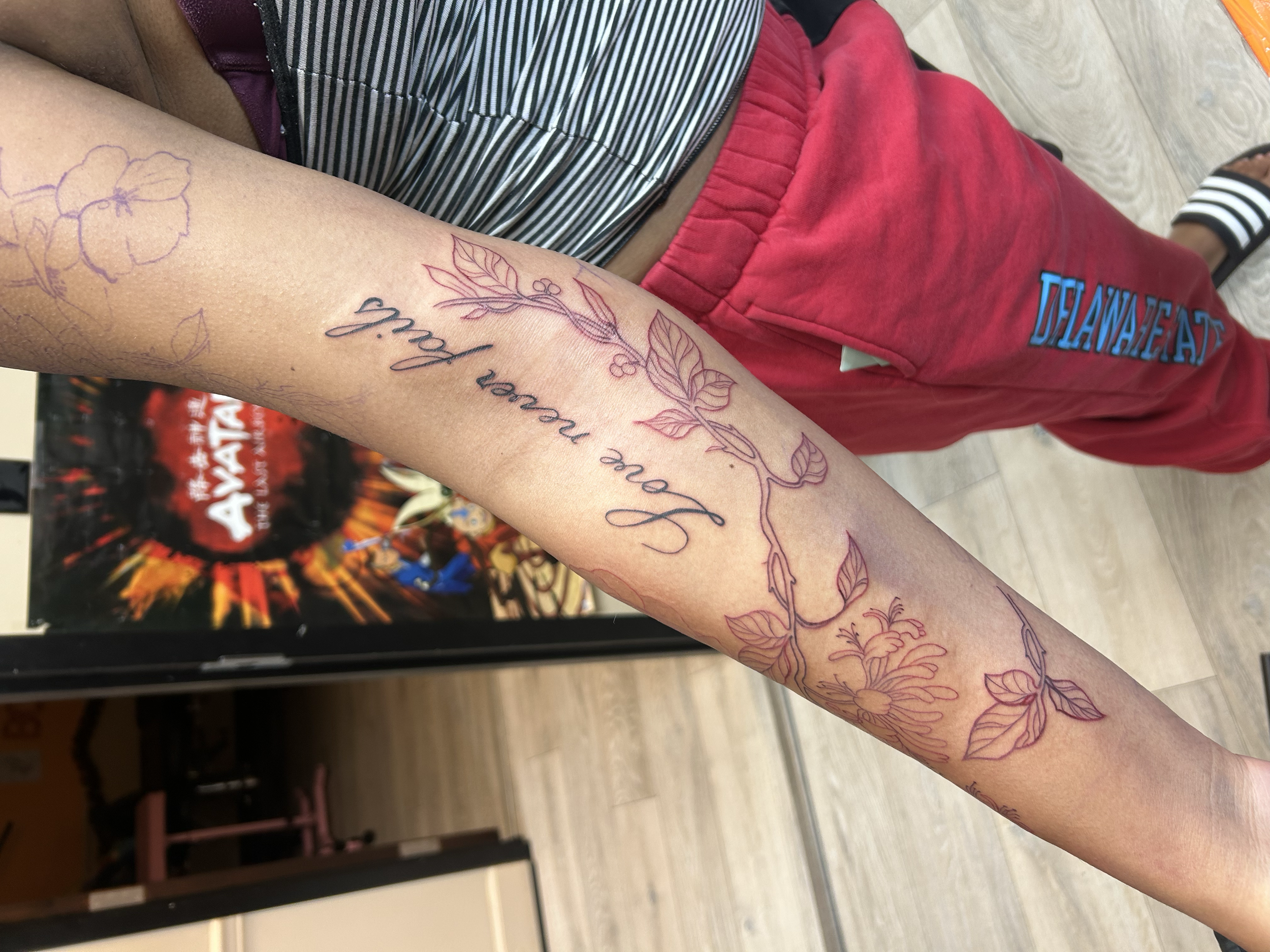 Close-up of a person's arm with multiple tattoos, including the phrases 'Love never fails' and various floral designs. The person is wearing red pants with blue lettering and striped slides. In the background, there is a vending machine and a movie p