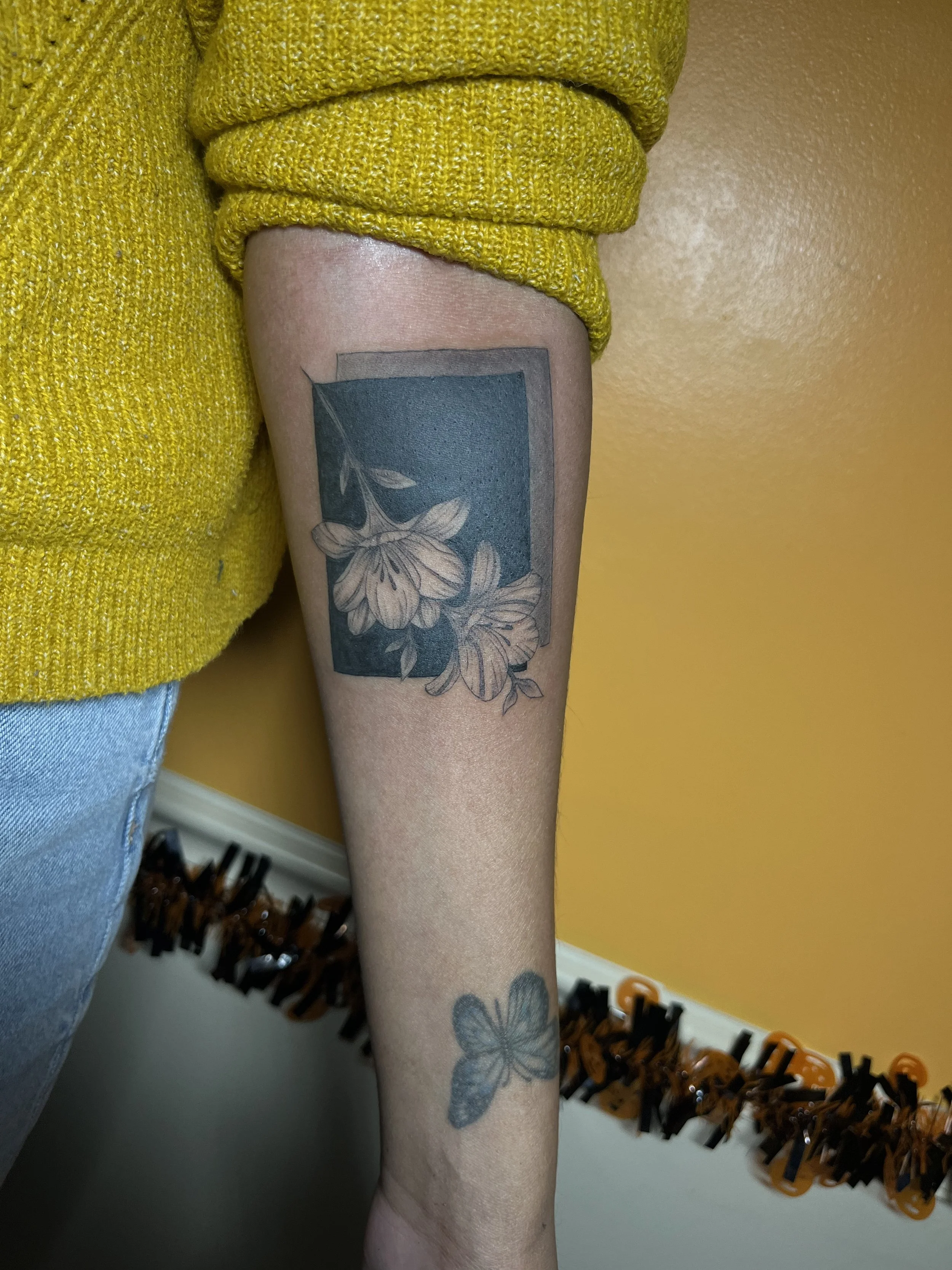Tattoo of a butterfly on the forearm below a rectangular frame with flowers inside, on a person's arm, with a yellow sweater and light blue jeans.