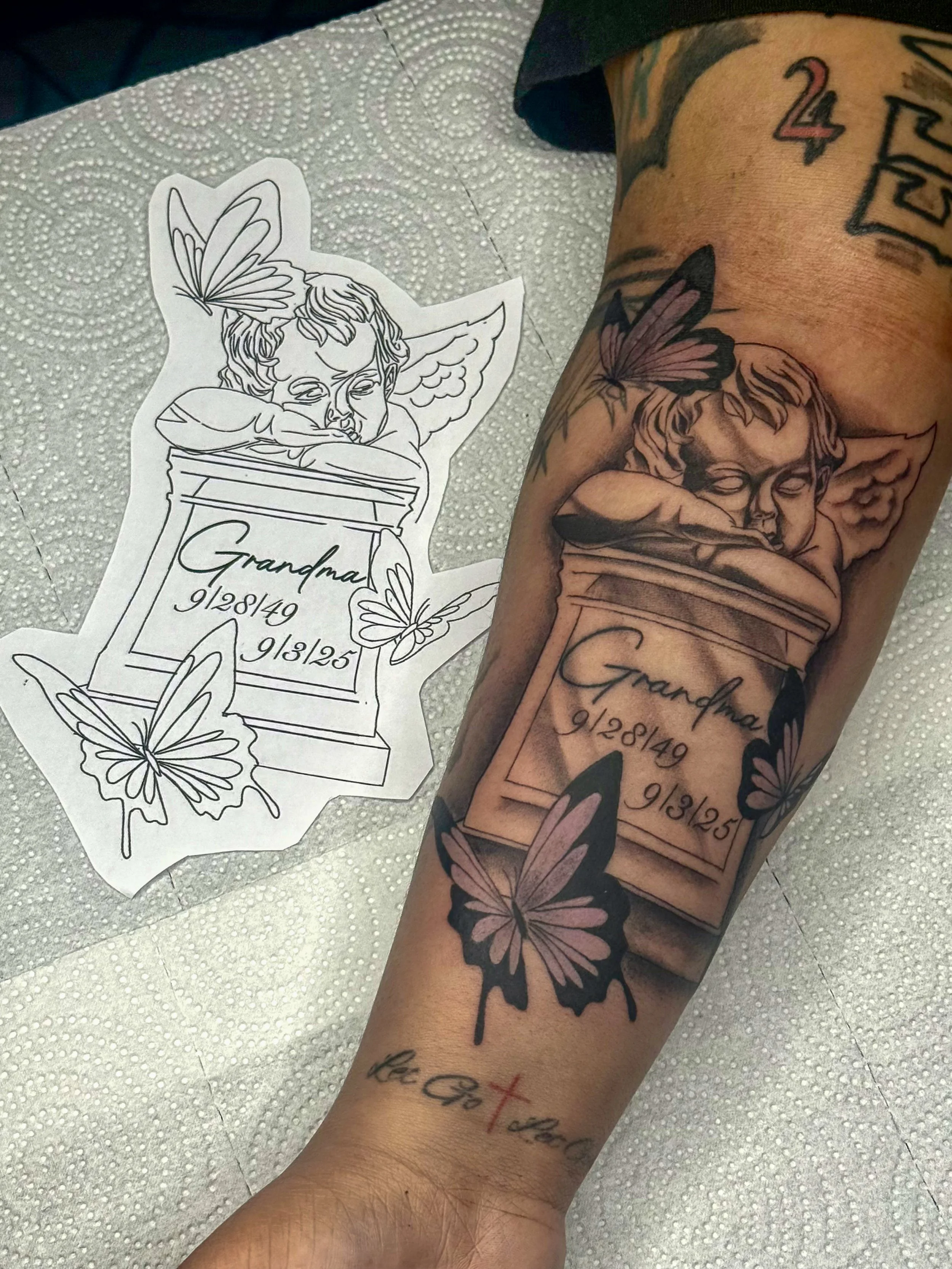 A tattoo on a person's forearm features a cherub with closed eyes resting its head on a memorial stone inscribed with the name "Grandma" and dates "9/28/49" and "9/3/25." The tattoo includes roses and butterflies surrounding the cherub and inscriptio