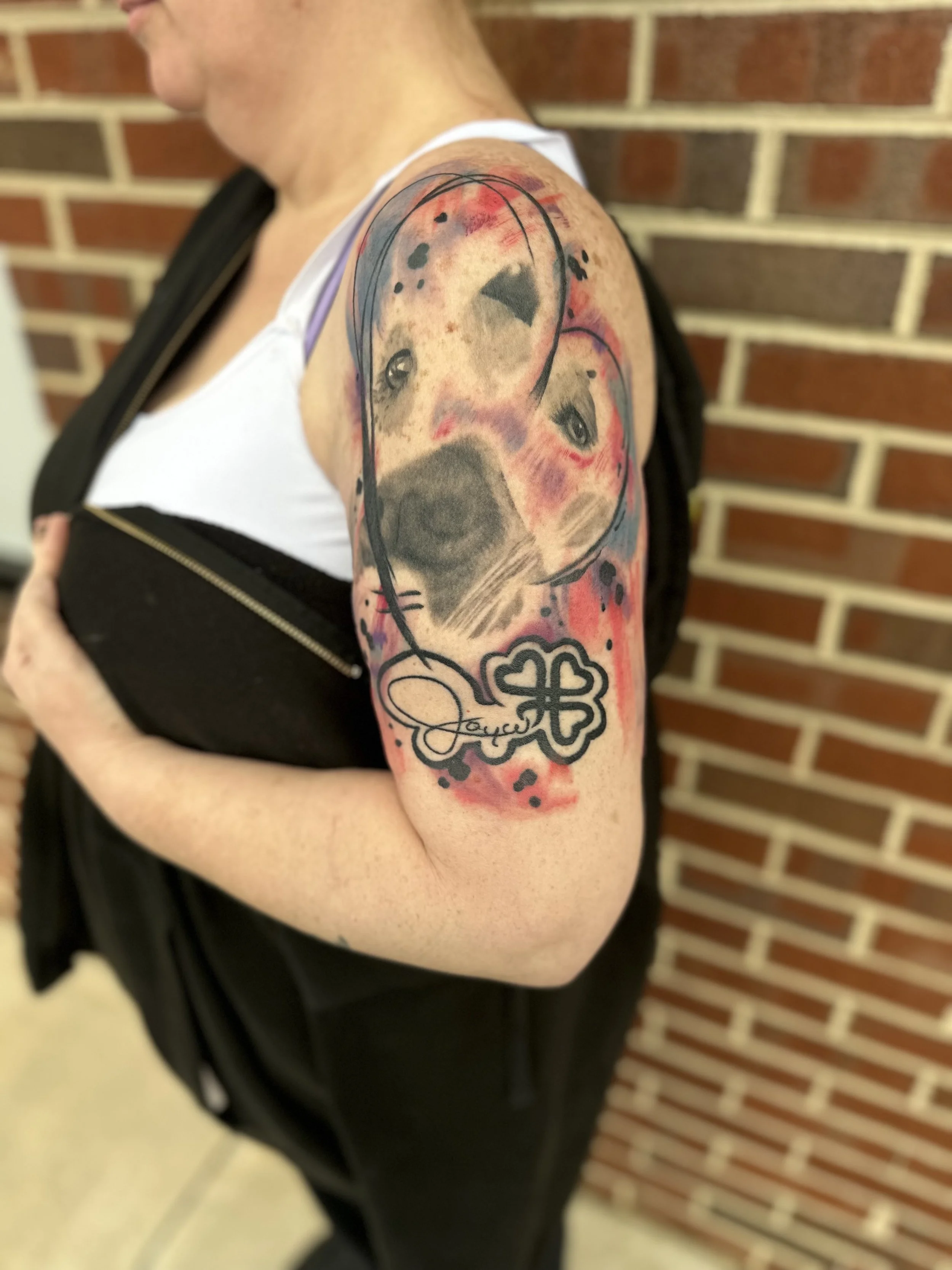 Tattoo of a dog's face with an abstract watercolor background on a person's upper arm, with the dog's name 'Joyce' in a decorative script.