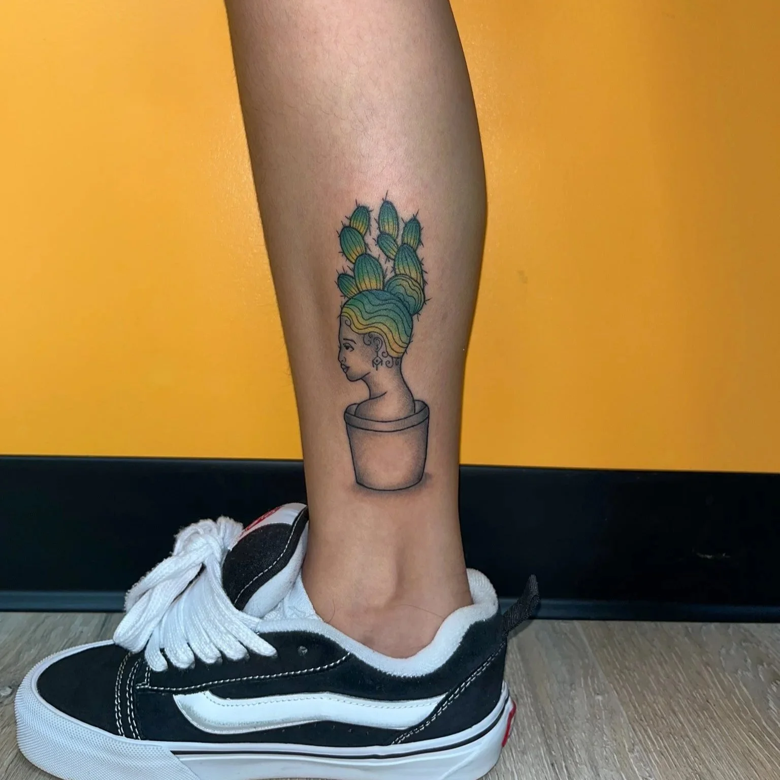 Tattoo of a woman's head with hair forming cactus plants in a pot, on a person's lower leg, with a black sneaker below