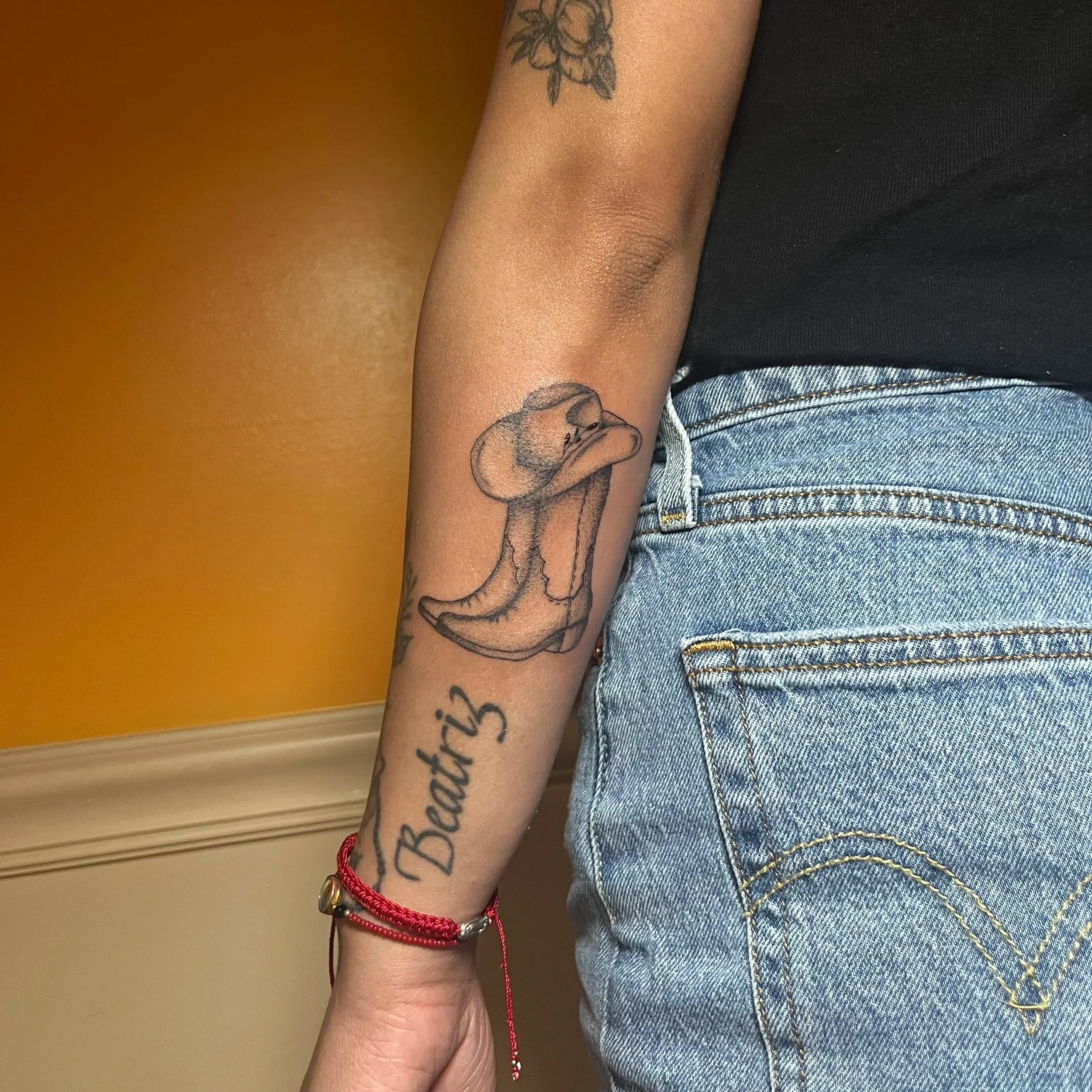 A person's arm with a tattoo of a bird and a tattooed word 'Beautiz'. The person is wearing a black shirt, blue jeans, and a red bracelet. The background is a yellow wall.