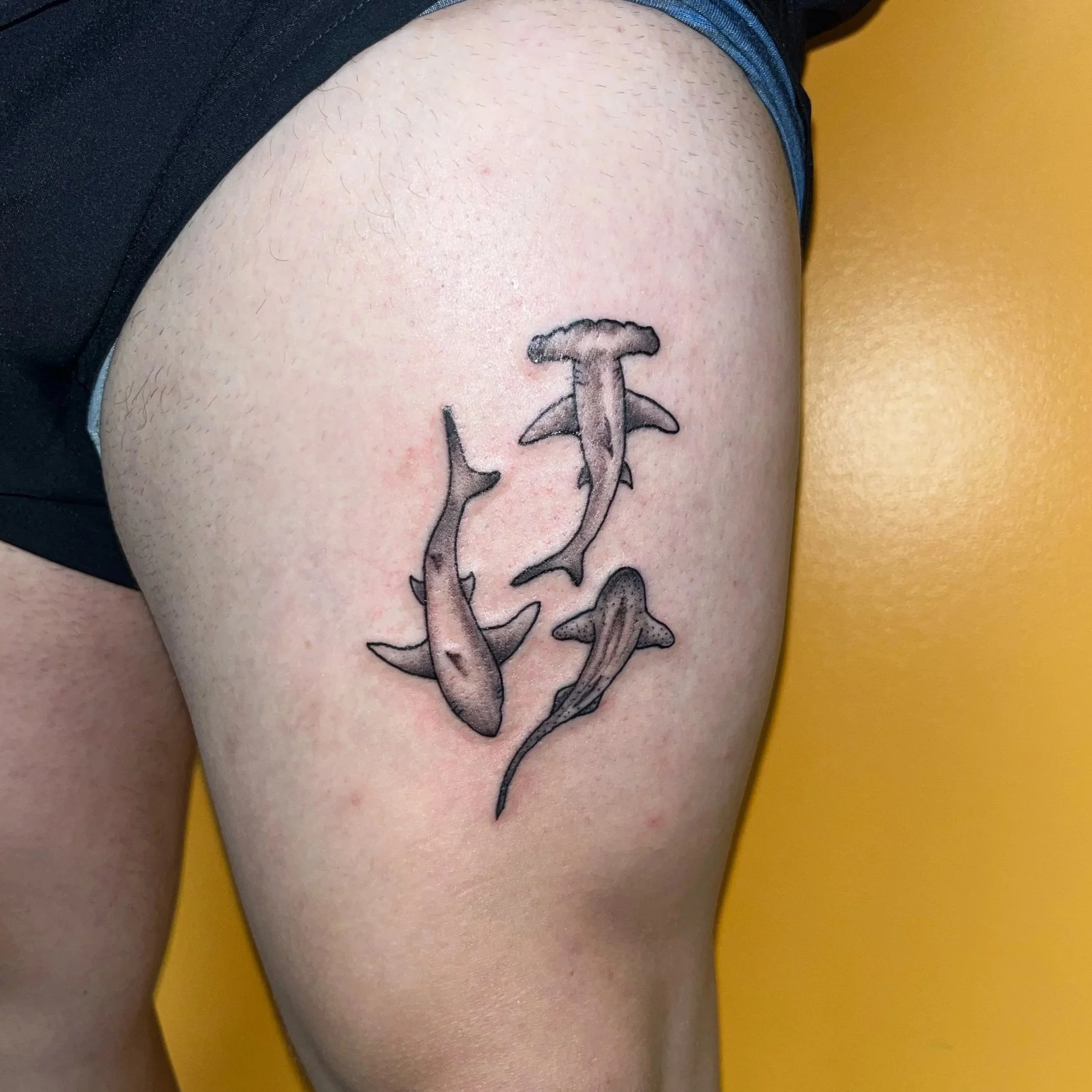 Tattoo of three sharks on a person's upper thigh, with the sharks in different positions and sizes, in a shaded black and gray style.