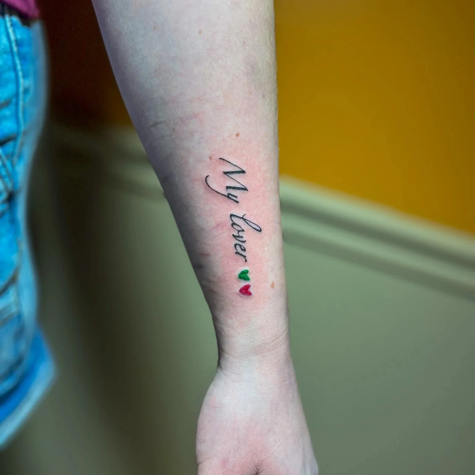 Tattoo on forearm with the words 'My Baby' in cursive, accompanied by two small hearts, one green and one red.