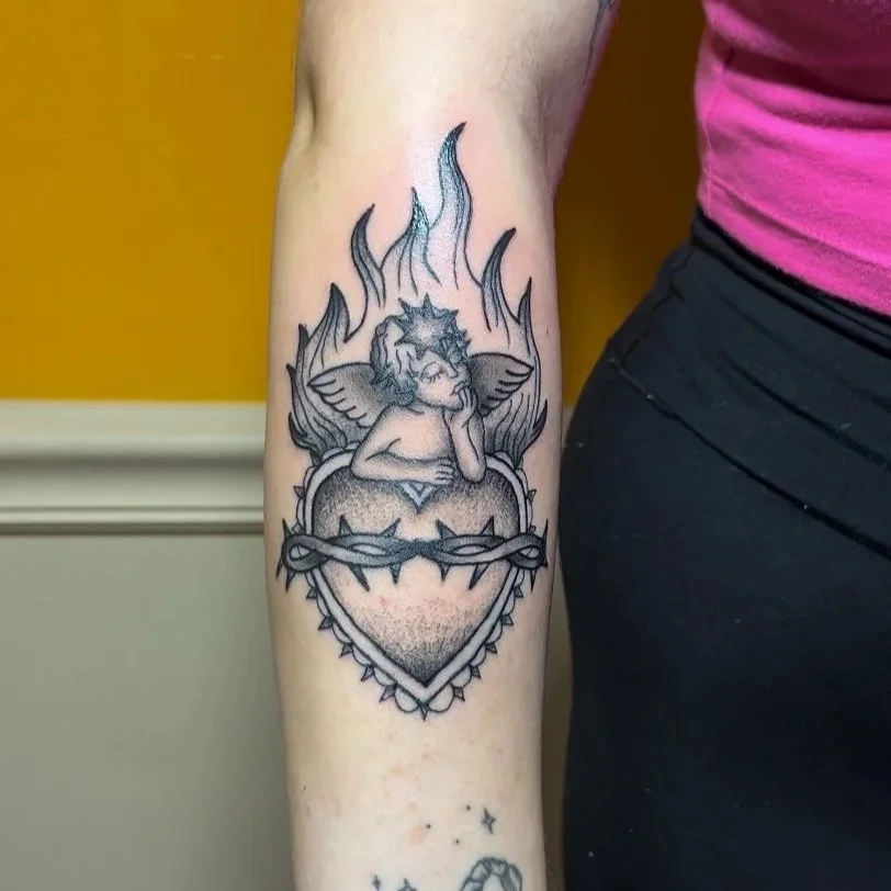 Tattoo of a angel with wings resting on a heart wrapped with barbed wire, surrounded by flames.