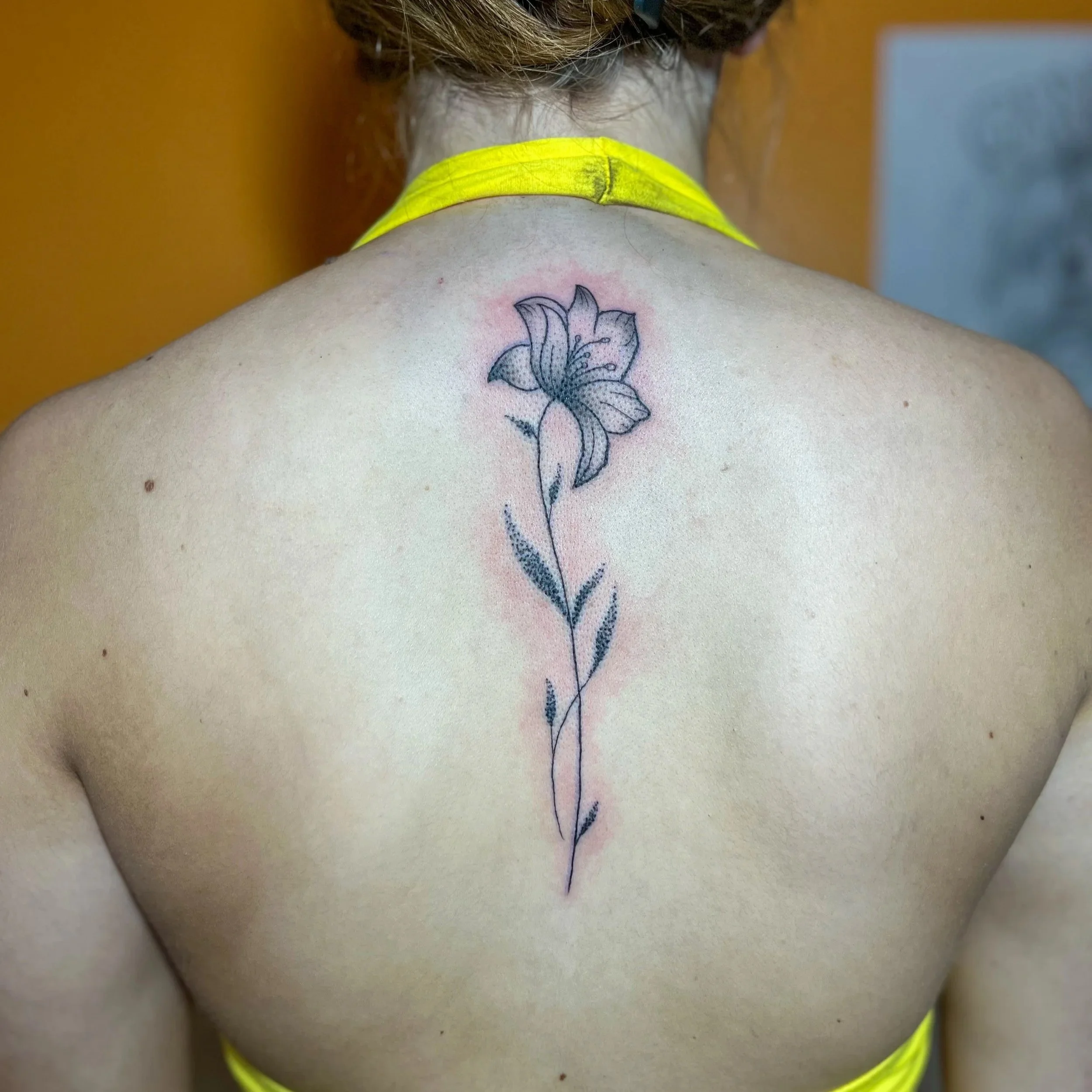 Tattoo of a lily flower in black and gray ink on a person's upper back, with a yellow strap visible around the neck.