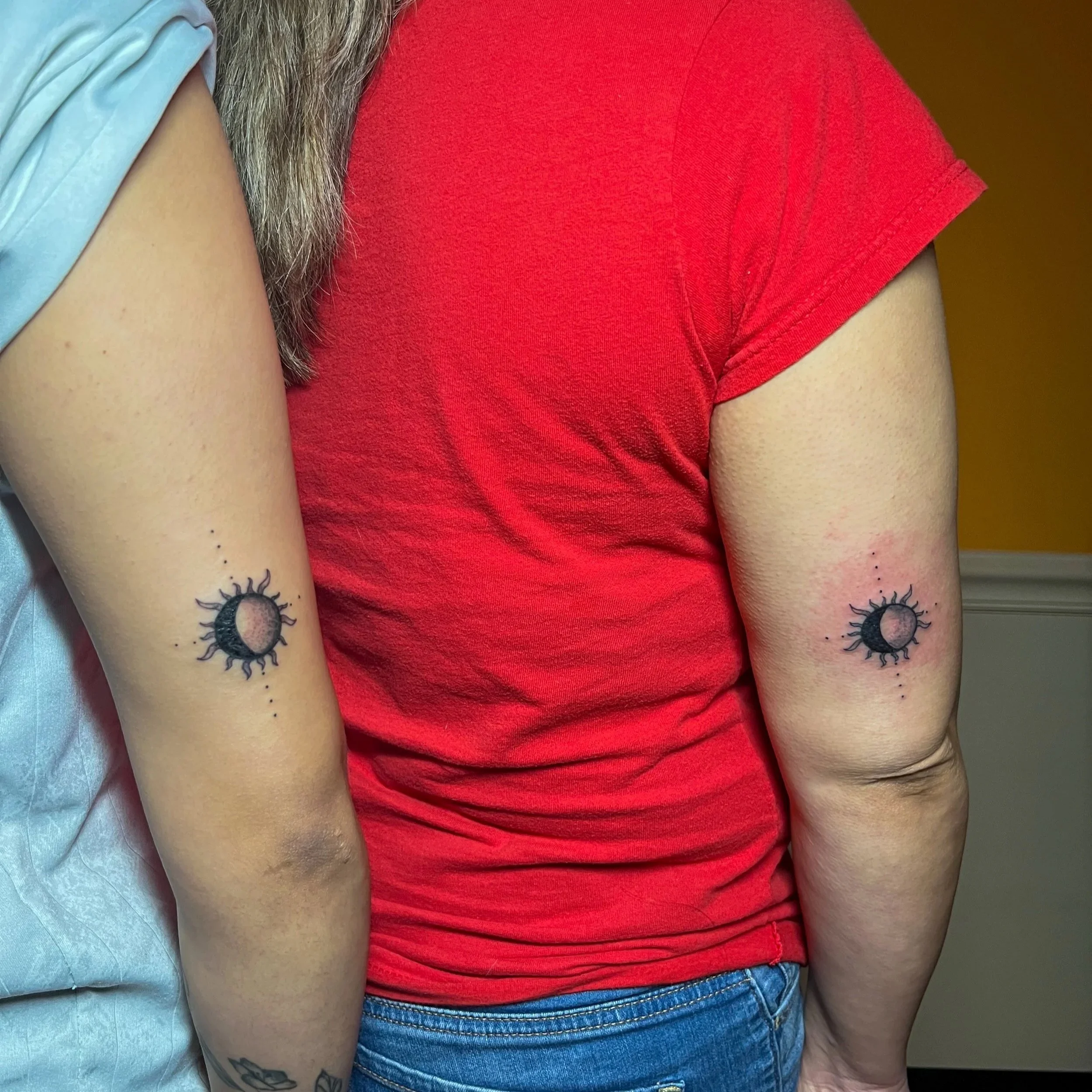 Two people with matching moon phase tattoos on their upper arms, featuring black moon symbols surrounded by small dots and lines.