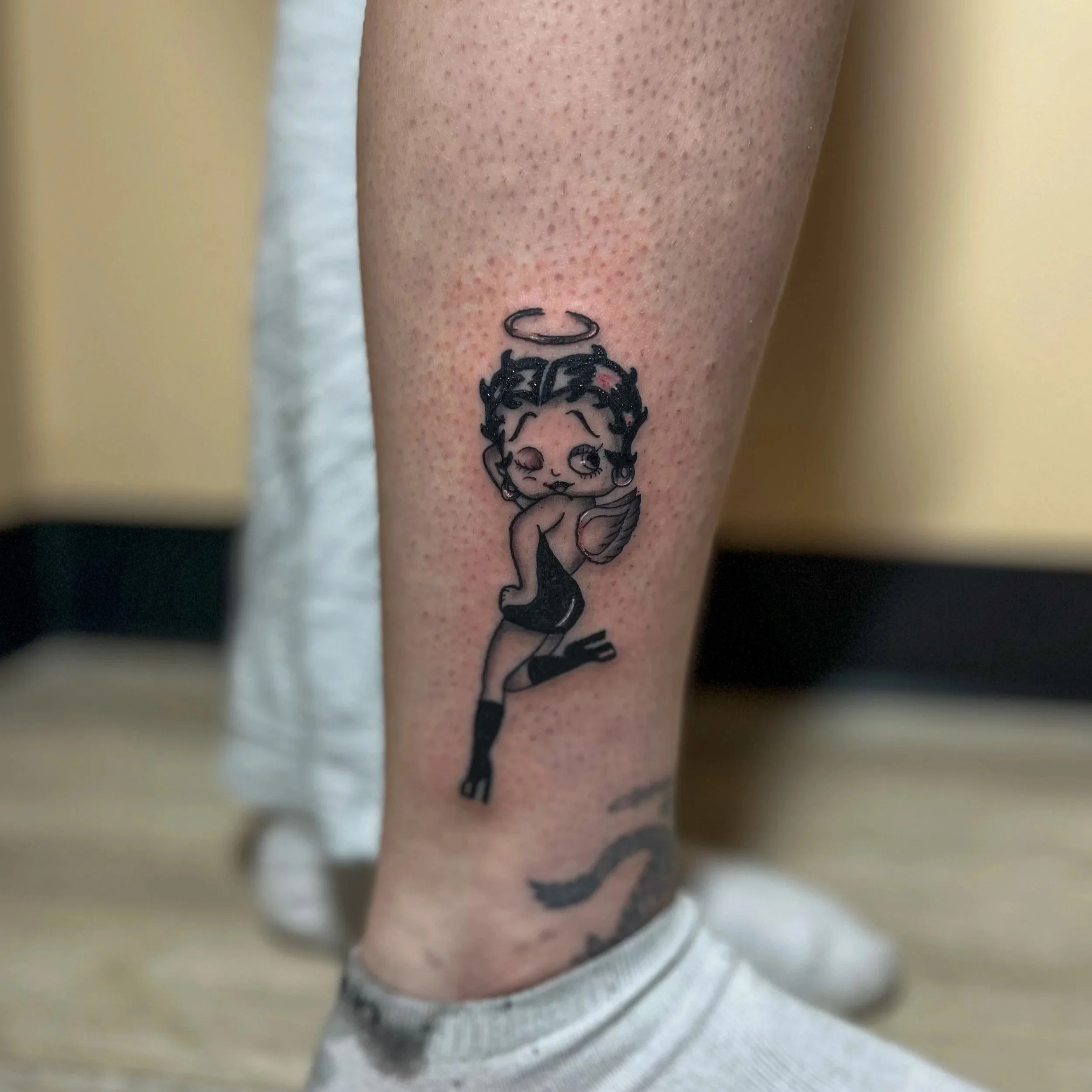 Tattoo of a cartoon angel with a halo, curly hair, and wings, winking and standing on one leg wearing a dress and high heels, on a person's calf.