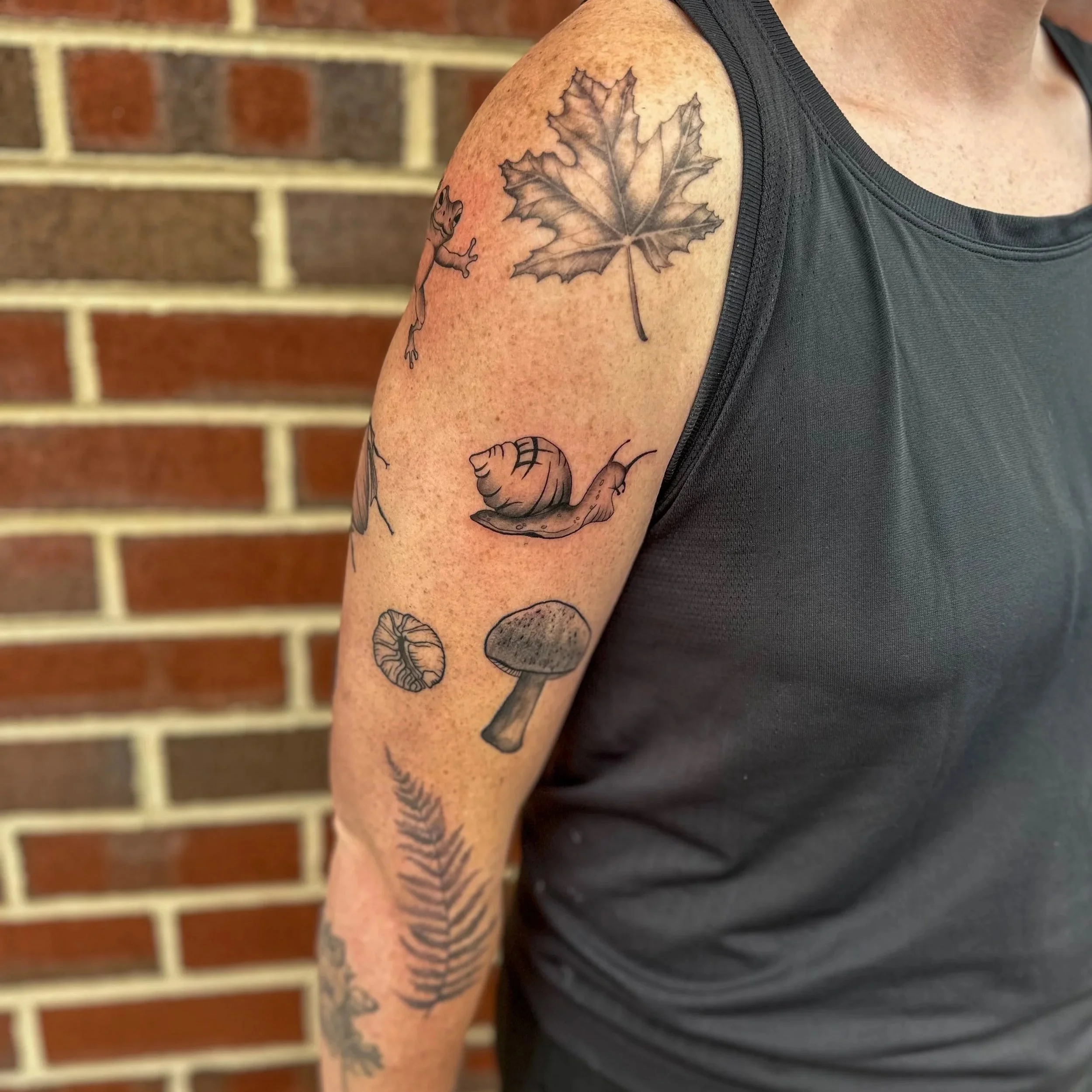 A person's upper arm with tattoos of a maple leaf, a snail, a mushroom, a fern, and a small animal.