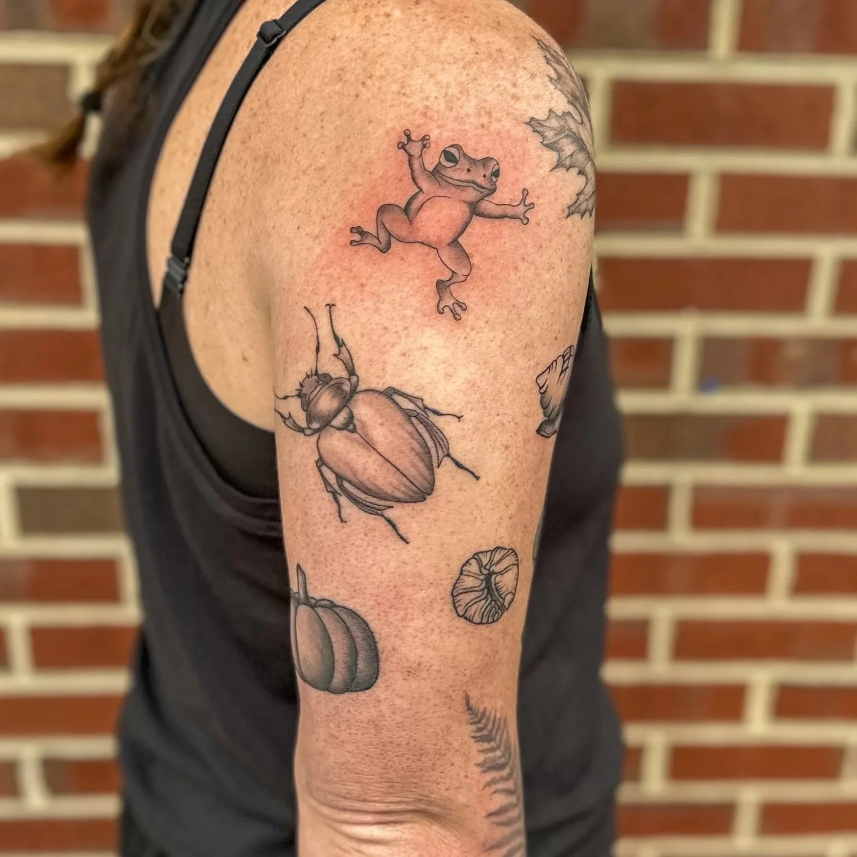 Tattoo of a frog on the upper arm, surrounded by various insects and leaves, with a brick wall in the background.