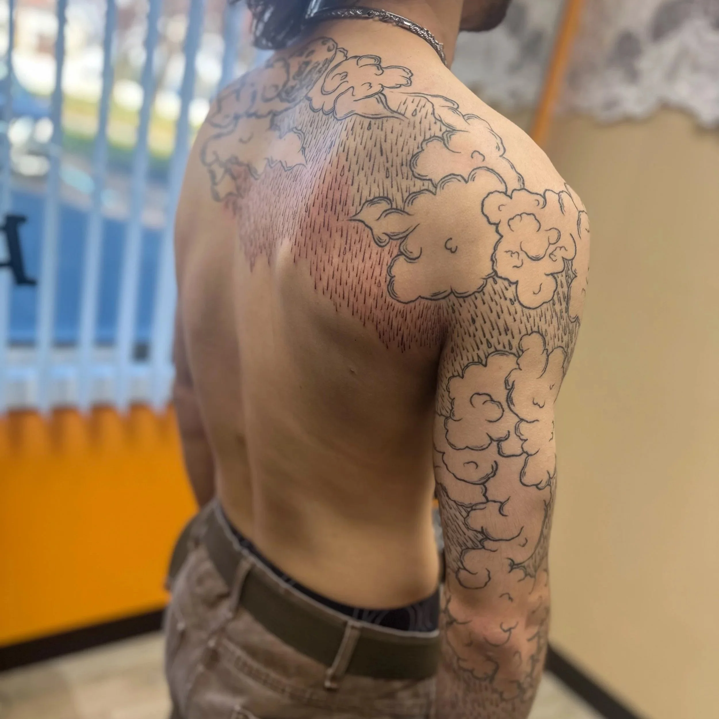 A shirtless man with tattoos of clouds and rain on his left shoulder and arm, wearing brown pants and a belt, standing indoors with a window and yellow wall in the background.