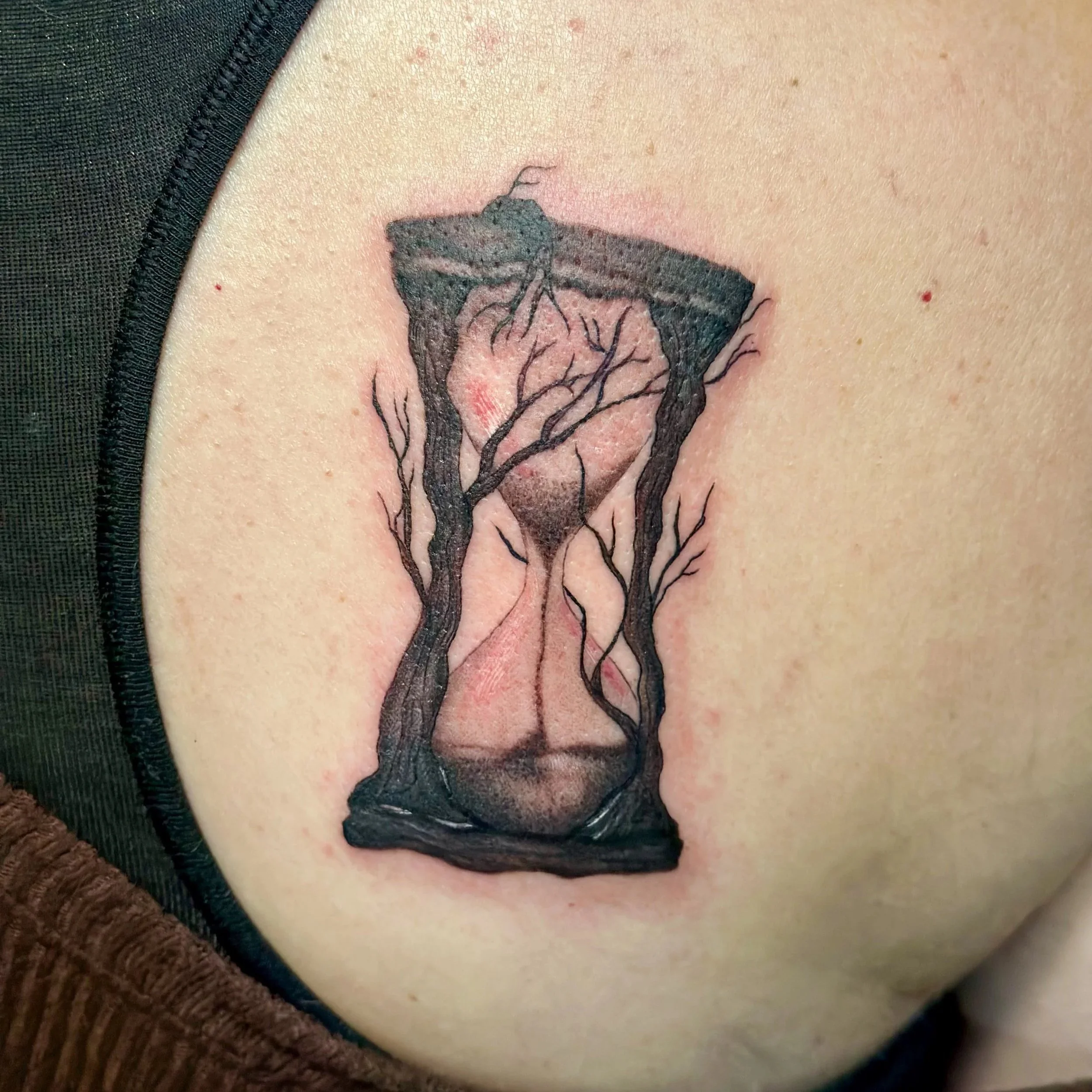 Tattoo of an hourglass with black tree branches intertwined around it on a person's shoulder.