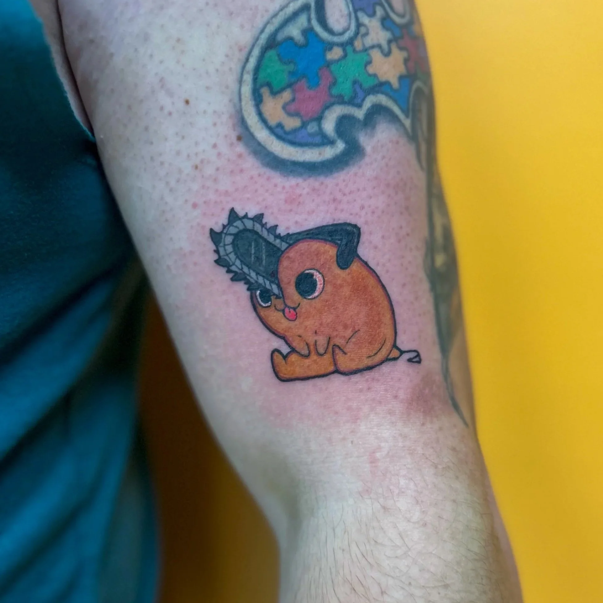Tattoo of a cute, cartoon-style character with large eyes and a small tongue sticking out, holding a chainsaw, on a person's upper arm.