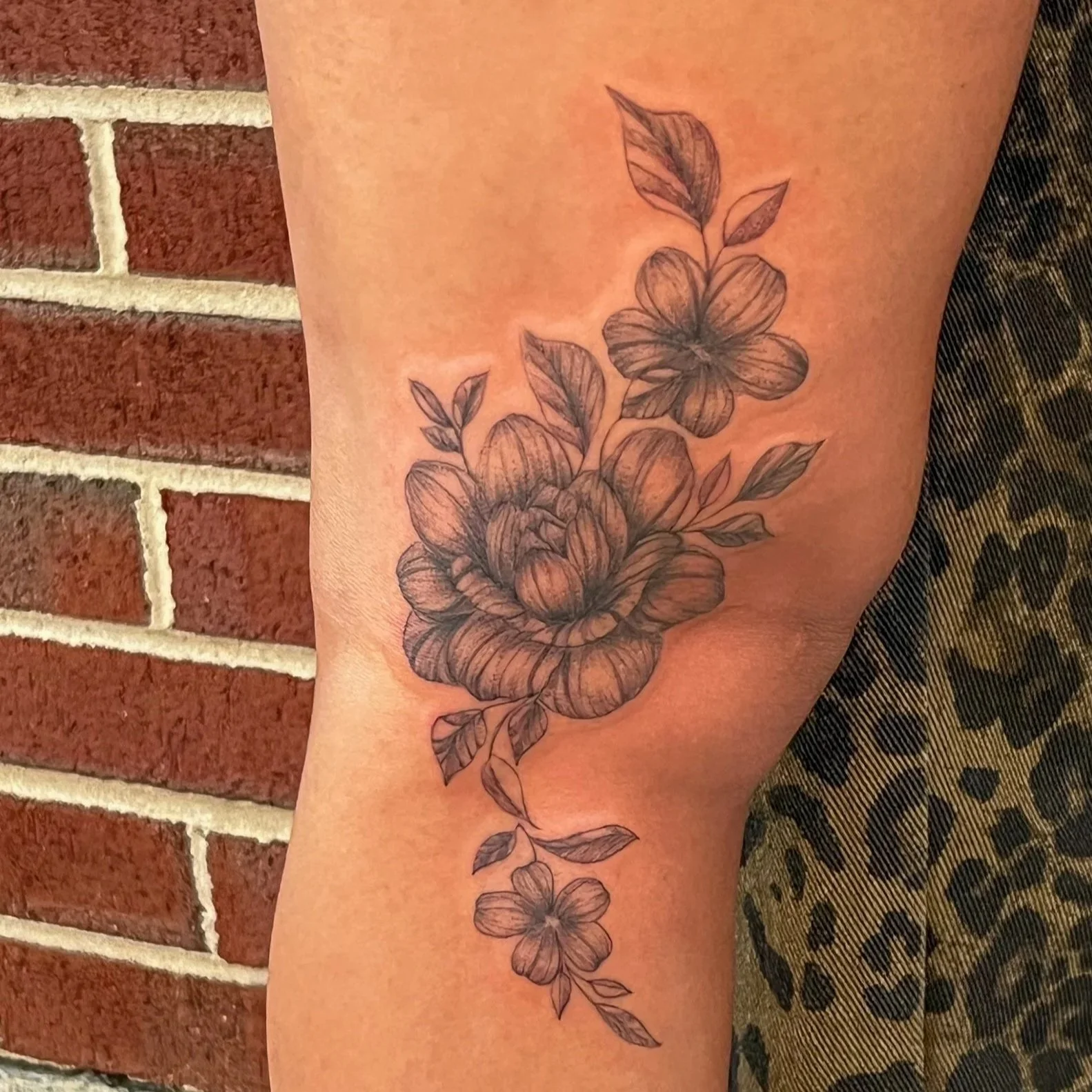 Black ink floral tattoo on person's arm, featuring a large blooming flower with detailed petals and leaves extending downward.