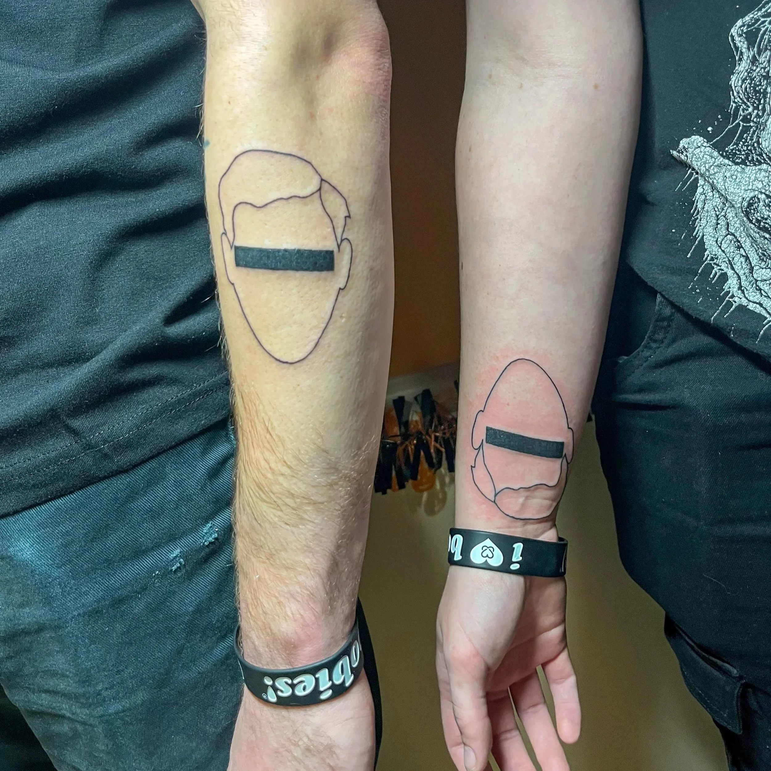 Two people with matching outline tattoos of faces with black bars over the eyes on their forearms. They are wearing black wristbands.