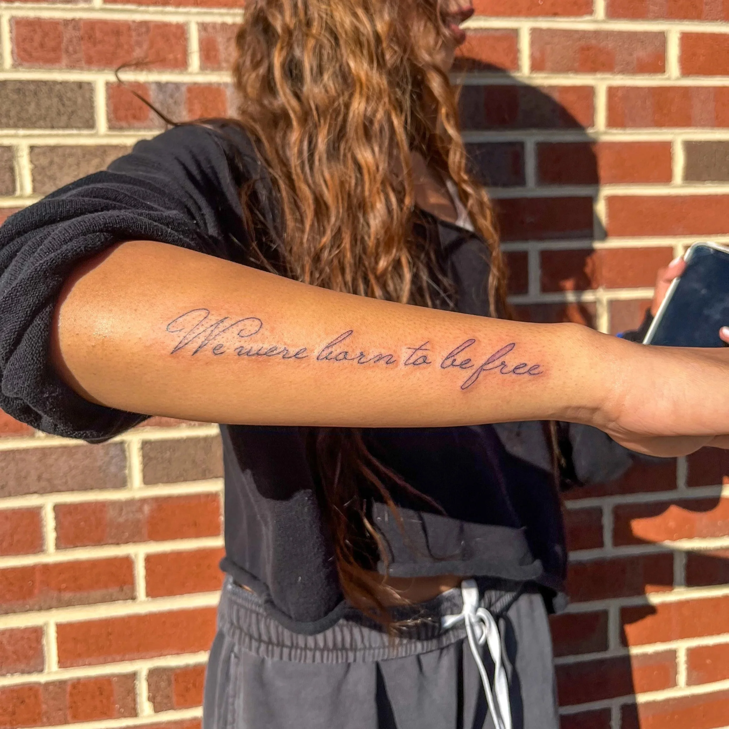 A person with long, curly hair holding a phone, showing a tattoo on their forearm that reads 'We were born to be free' in cursive script, with a brick wall in the background.