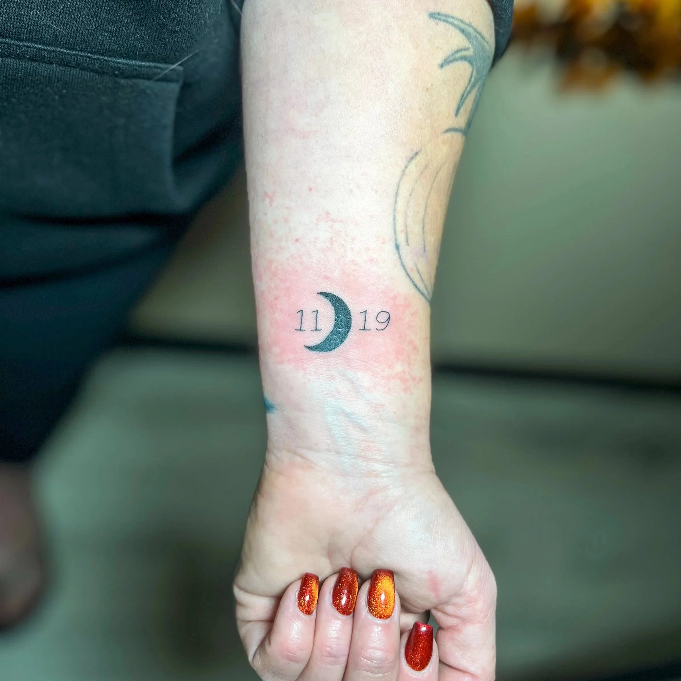Tattoo of a crescent moon with the numbers 11 and 19 on either side on a person's forearm.
