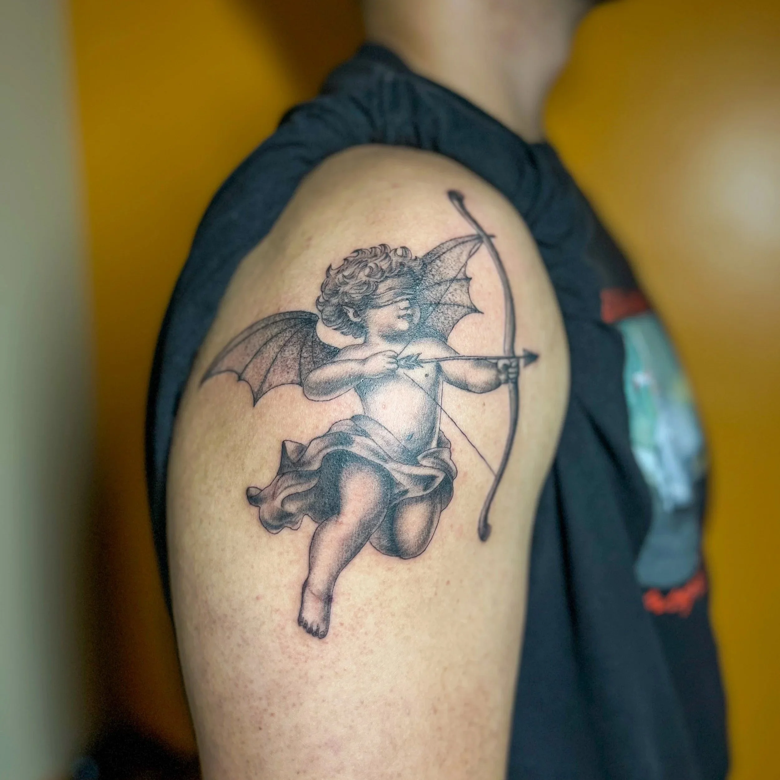 Tattoo of a cherub with bat-like wings drawing a bow, on a person's upper arm.