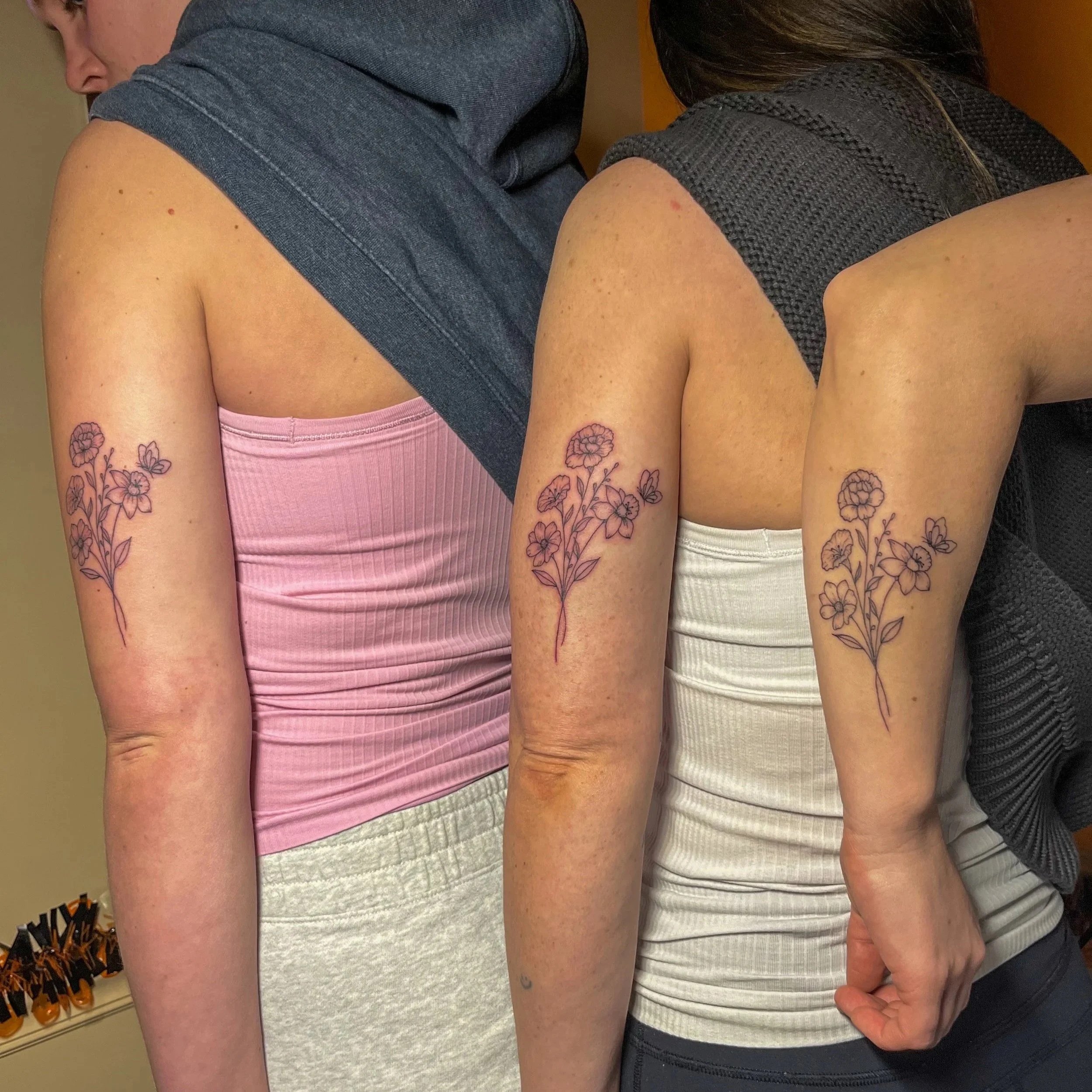 Two women stand side by side showing their tattoos of a flower branch on their upper arms.