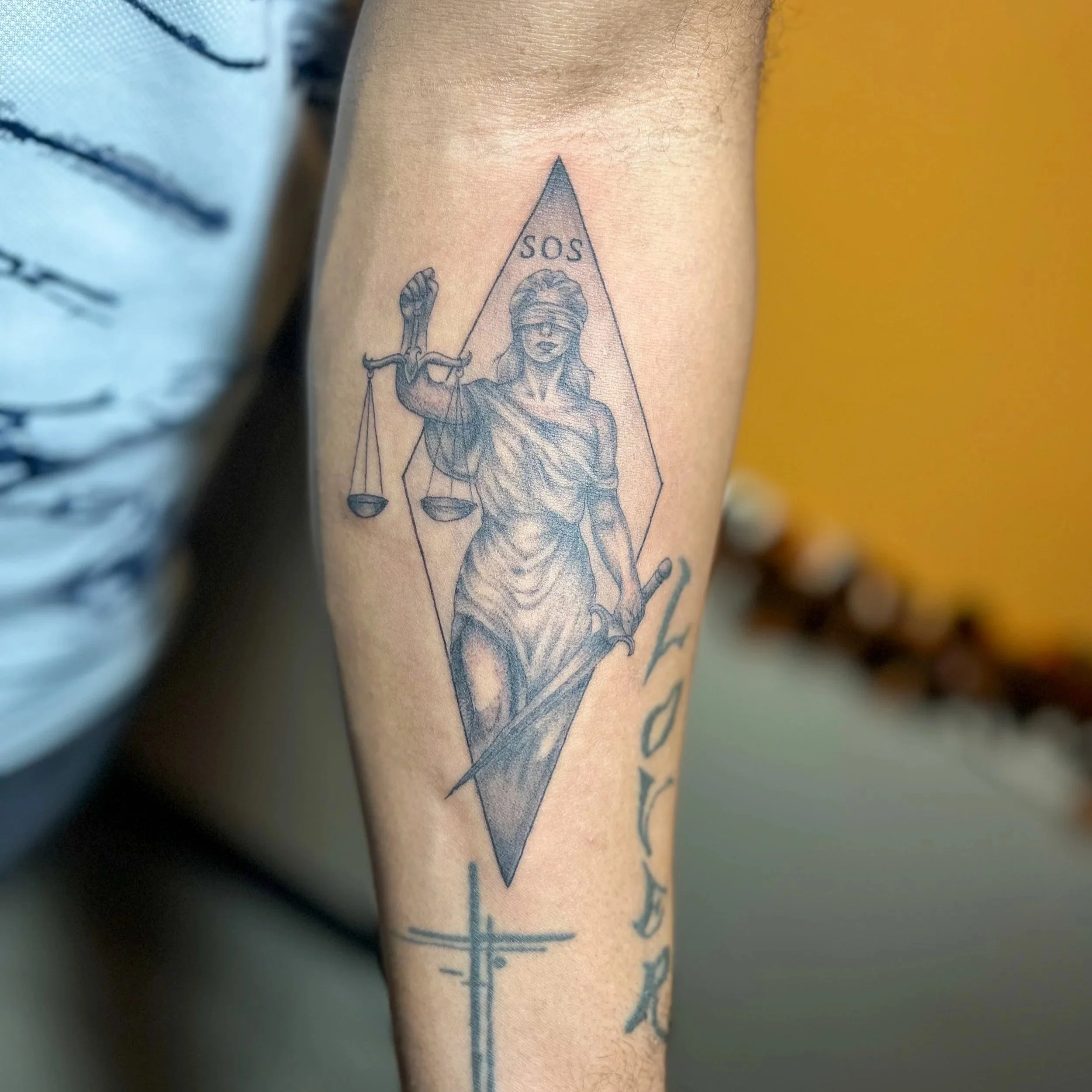 Tattoo of Lady Justice with a blindfold holding scales and a sword, with the letters "SOS" above her head, on a person's forearm.