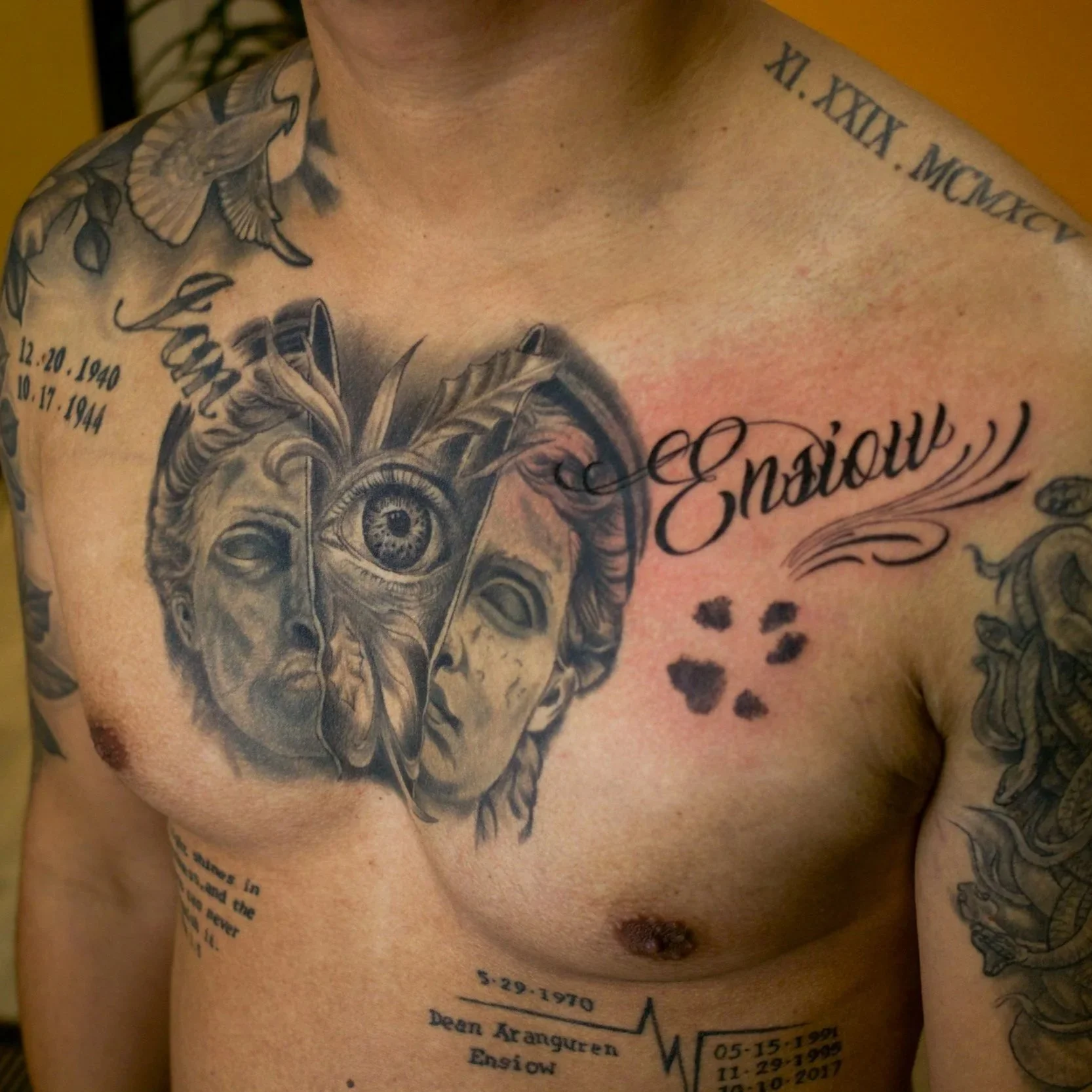 A person's chest tattoo featuring a classical sculpture face with an eye symbol and leaves, accompanied by the word "Ensiow" in cursive, floral elements, paw print, dates, and additional text.