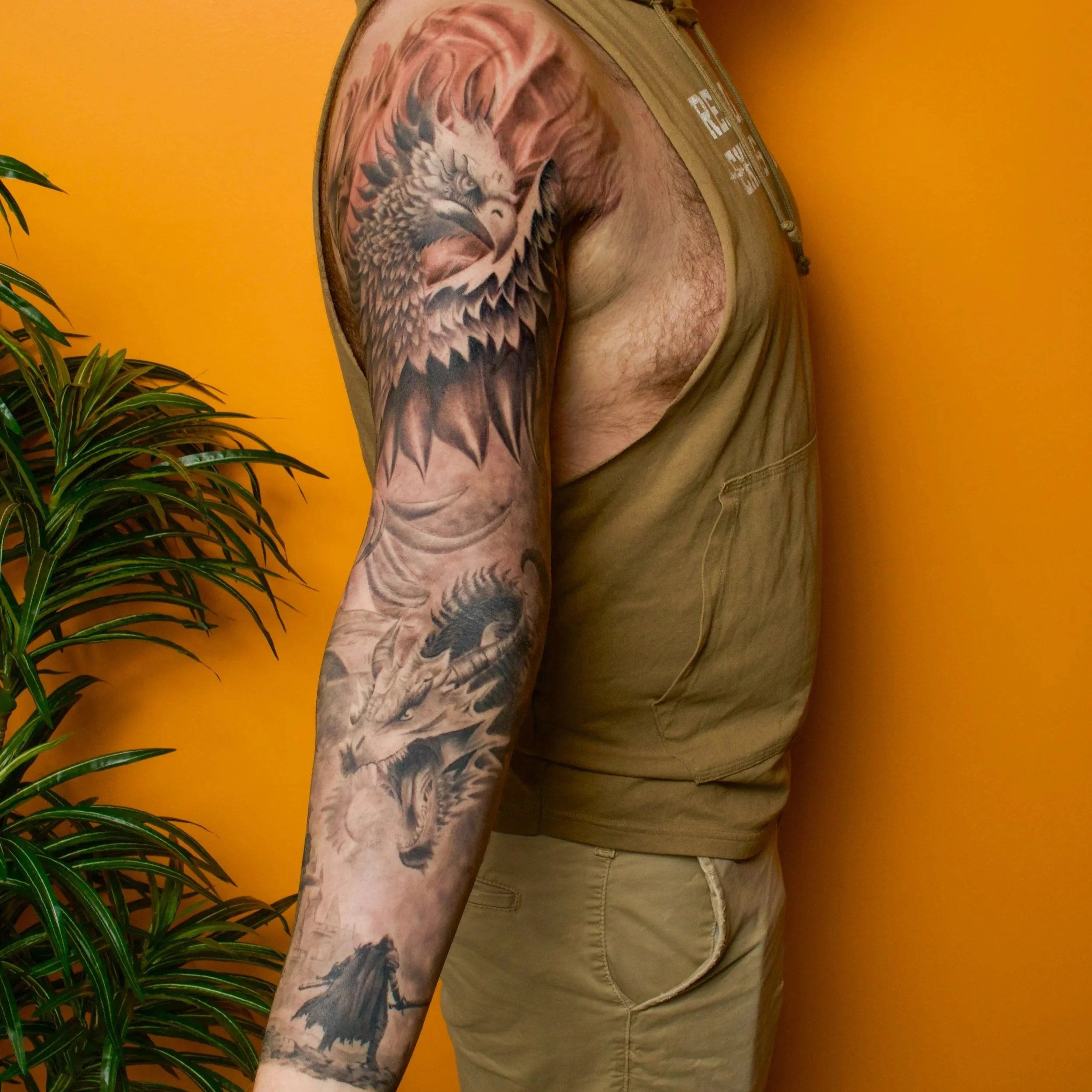 A person with a large tattoo sleeve on their arm, featuring a detailed pheonix and a fierce dragon with a solider holding a sword on their shoulder, standing against an orange background.