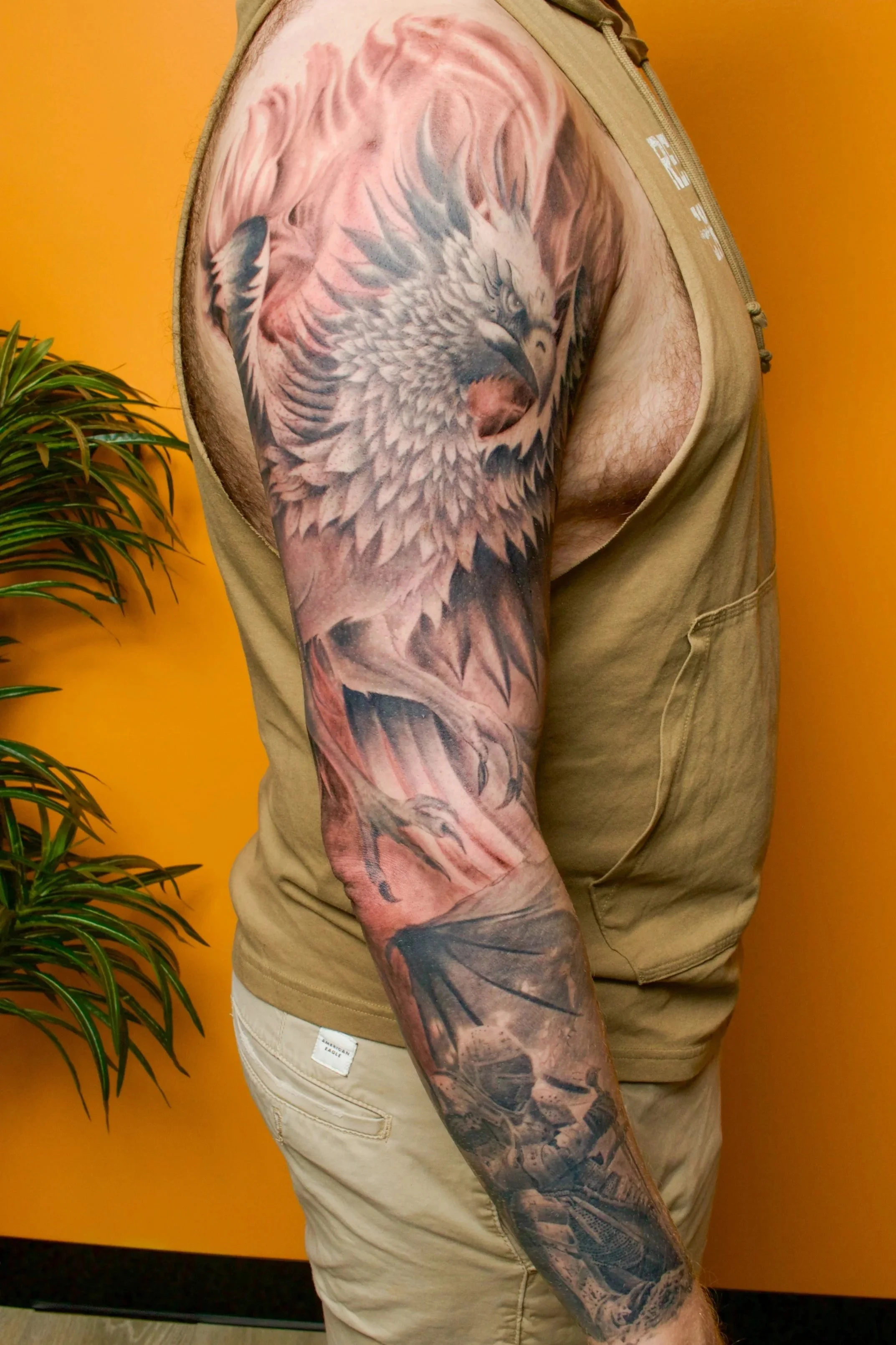 Tattoo of a fierce-looking phoenix with spread wings covering the person's upper arm and shoulder, against an orange wall background.
