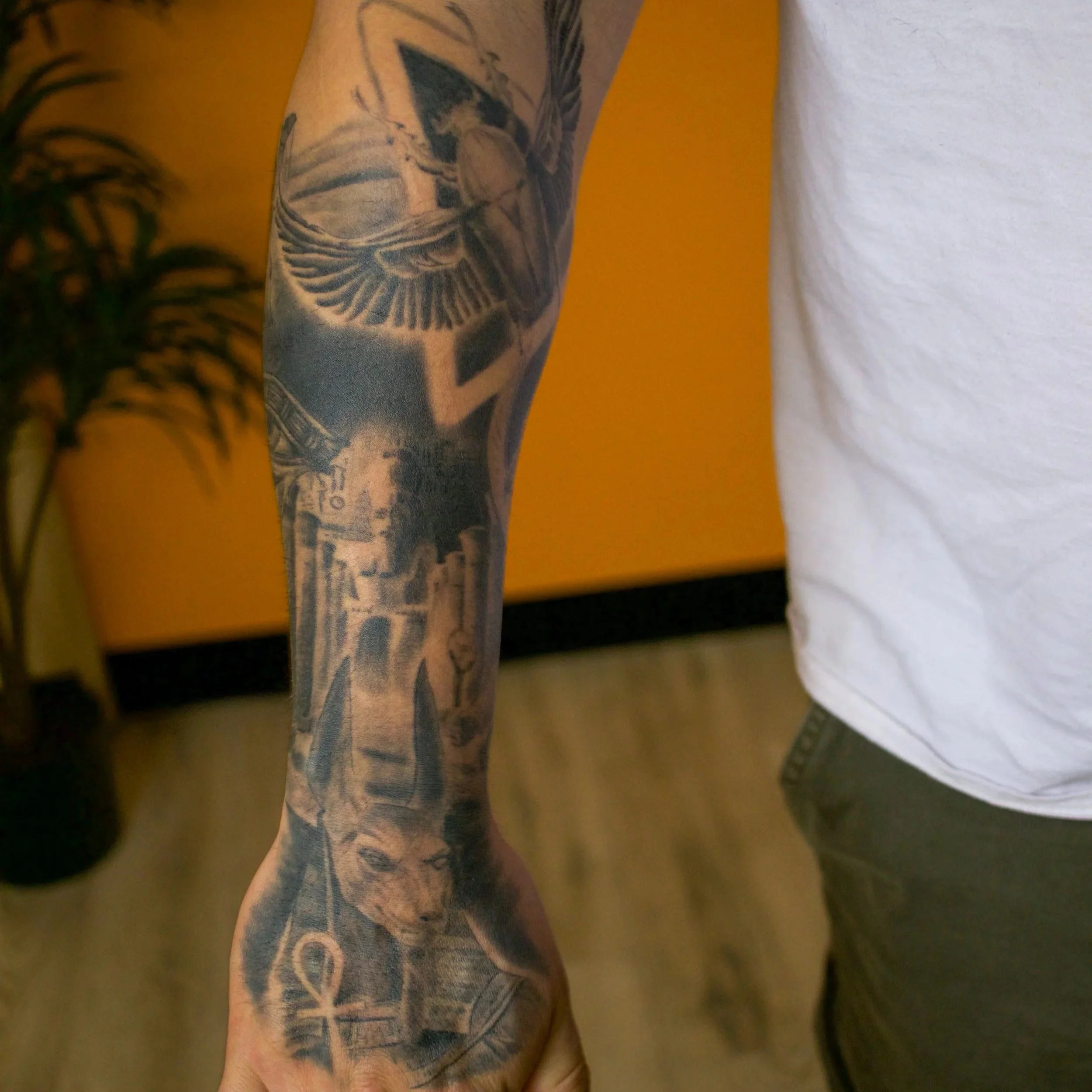 A person's arm with a detailed black and gray tattoo sleeve that includes a cat's face, Egyptian ankh symbol, and various other images.
