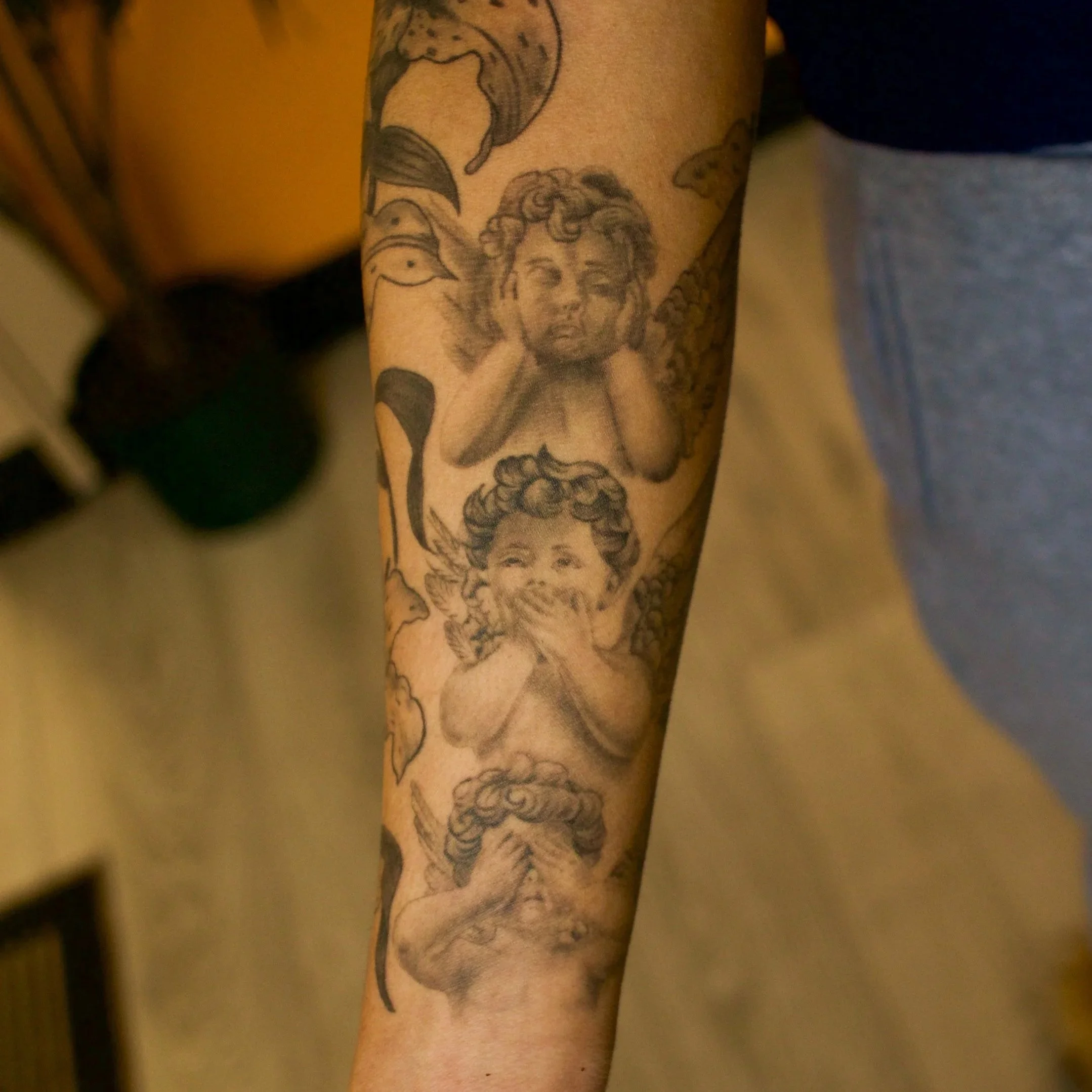 Black and grey tattoo of three cherubs on a person's forearm, with clouds and decorative elements.