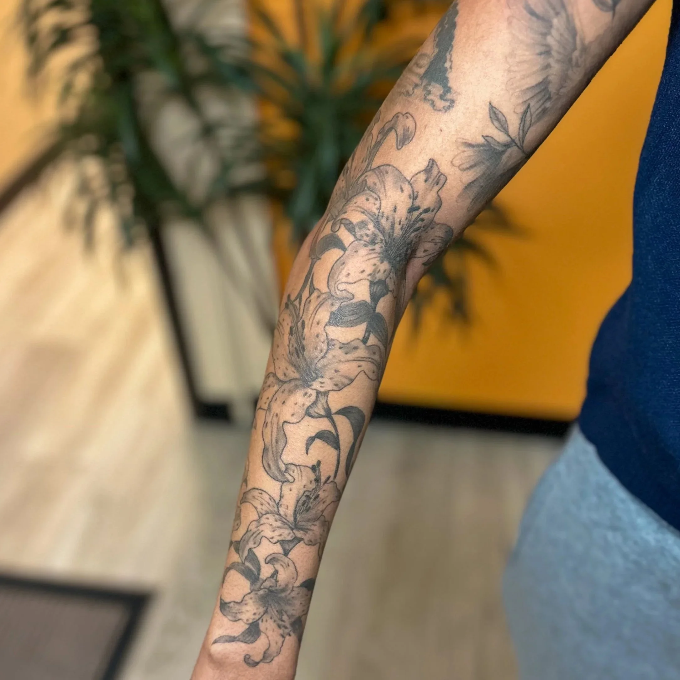 Close-up of a person's arm with a detailed black ink floral tattoo featuring lilies and leaves.