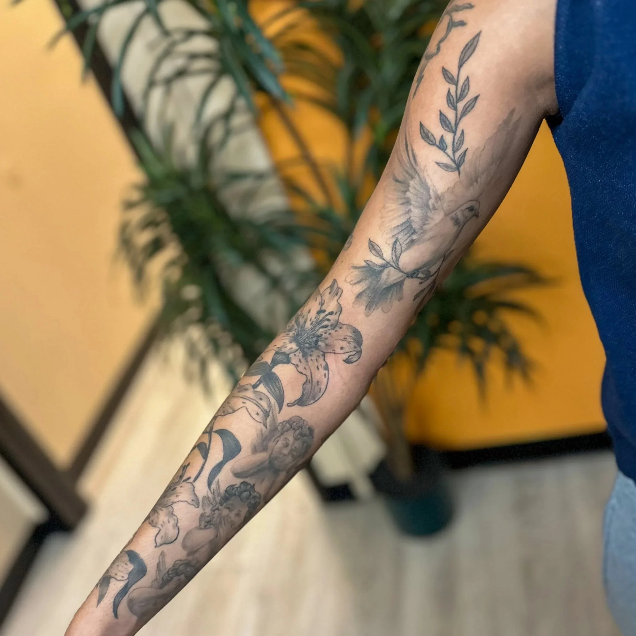 Tattooed arm with floral and bird designs, including a lily, a bird in flight, and a woman's face, against a background of indoor plants and yellow walls.