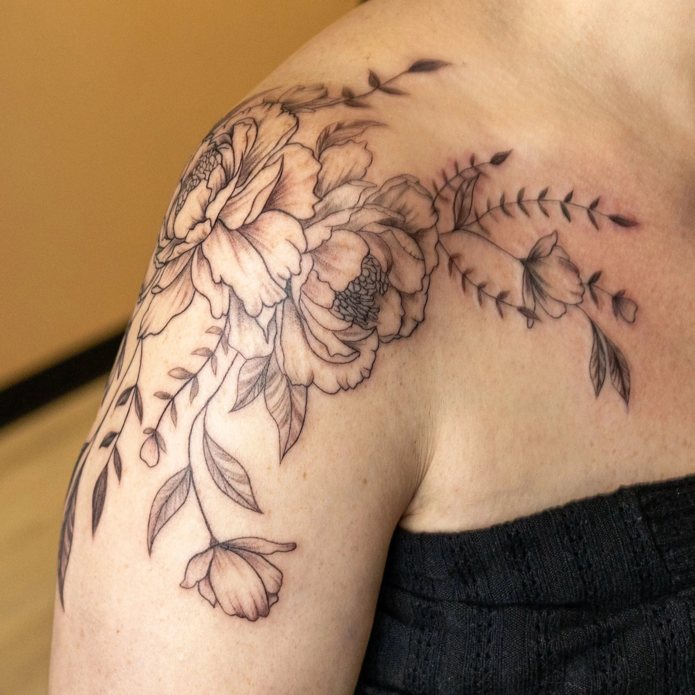 A partial view of a person's upper arm and shoulder showcasing a black ink floral tattoo with detailed petals and leaves.